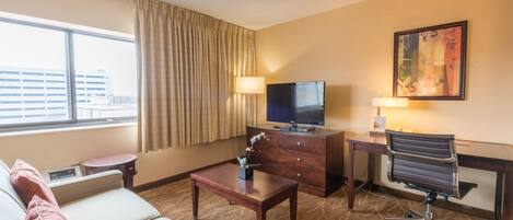 Une chambre d’hôtel équipée d’un canapé, d’un bureau avec un ordinateur, d’une télévision, d’une lampe et d’une fenêtre avec des rideaux.