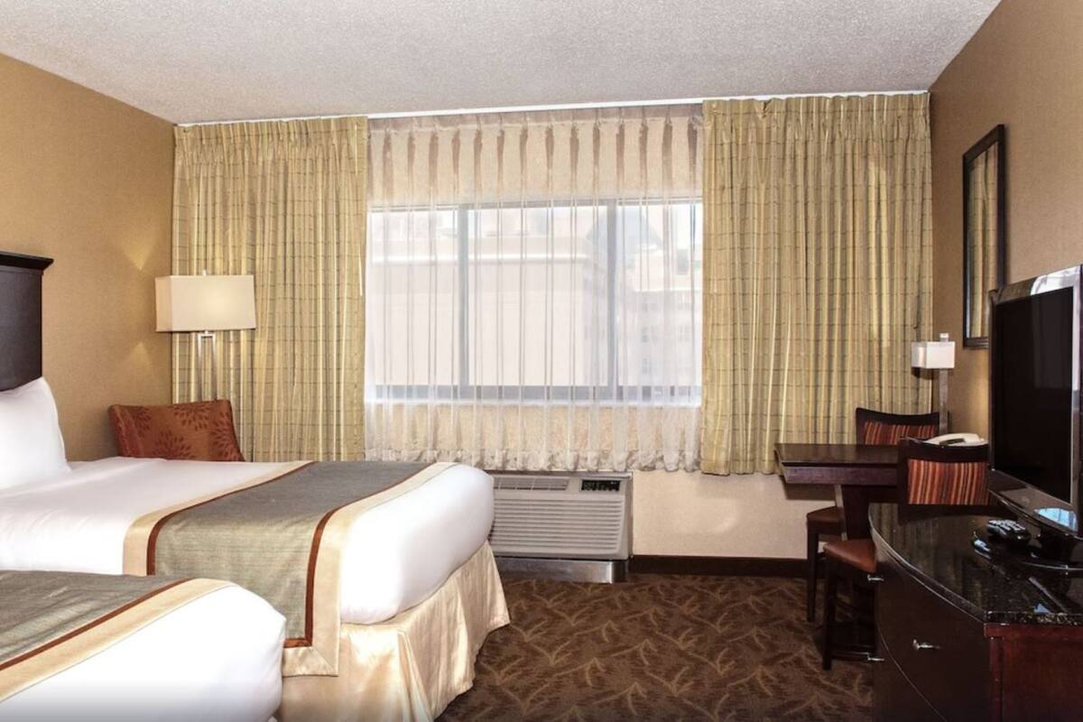 A hotel room with two beds, a desk with a TV, a lamp, and a window with curtains.