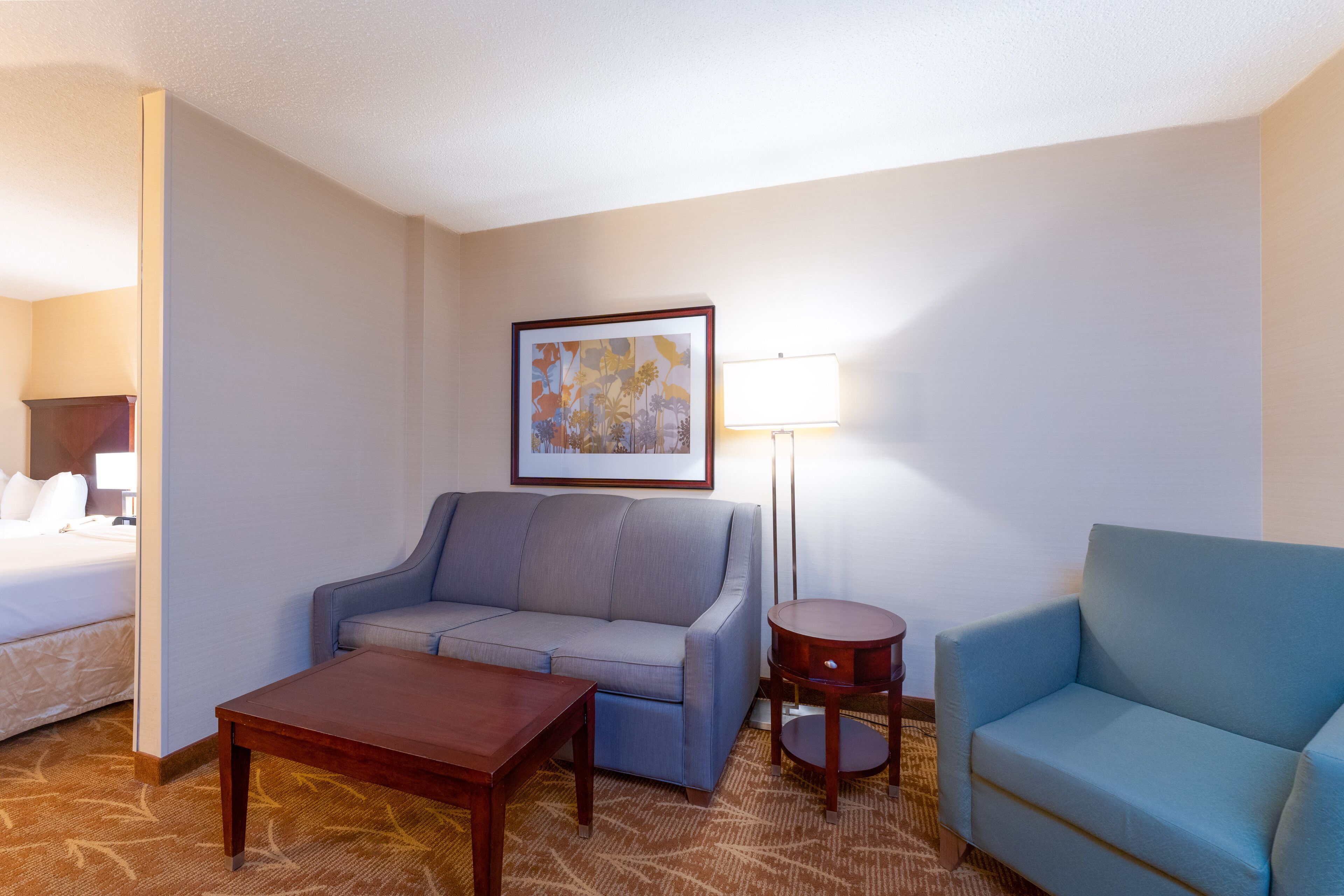 A hotel room with a bed, a grey sofa, a blue sofa, a wooden coffee table, a floor lamp, and a framed picture on the wall.