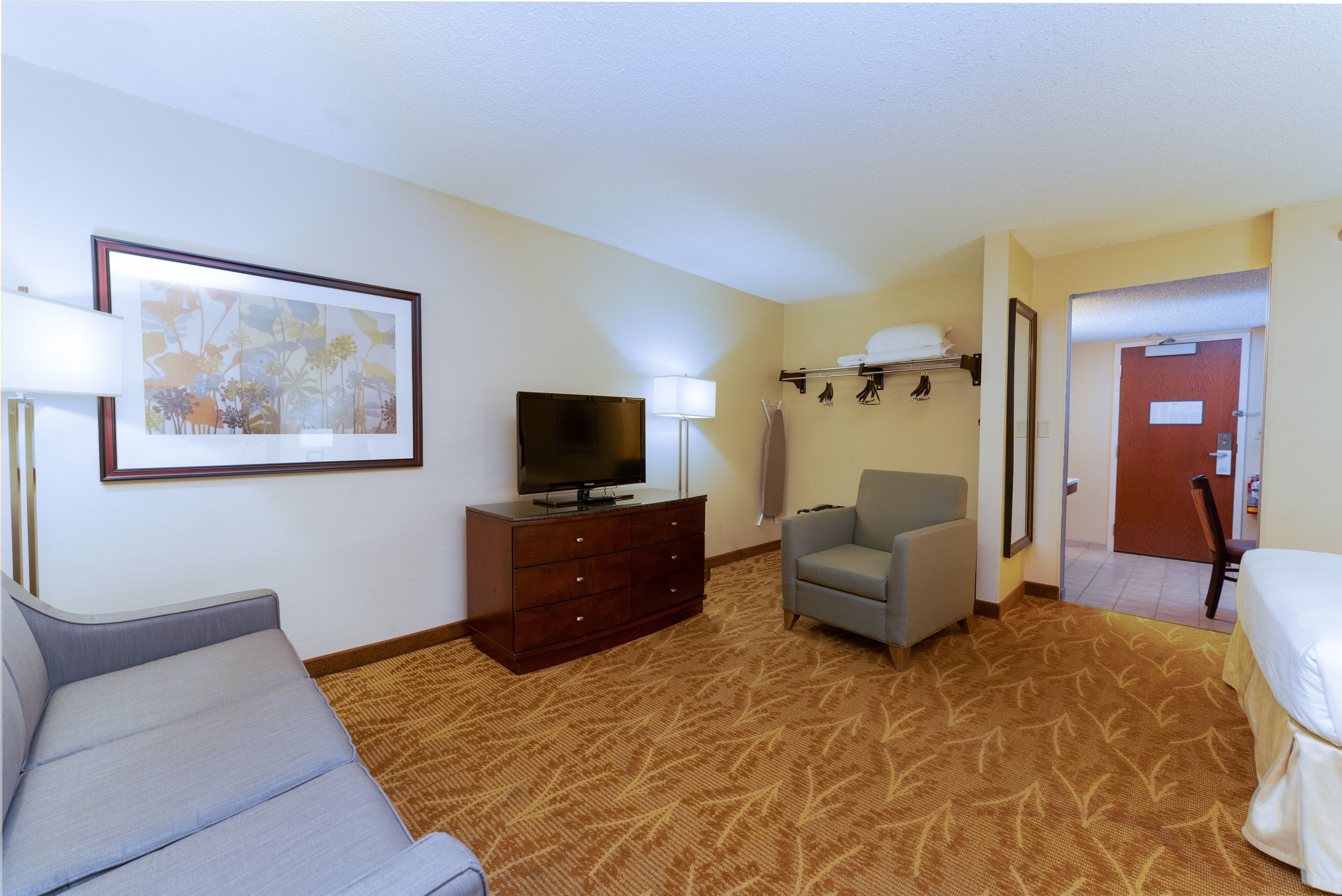 A hotel room with a sofa, a TV on a wooden dresser, a grey armchair, and a patterned carpet.