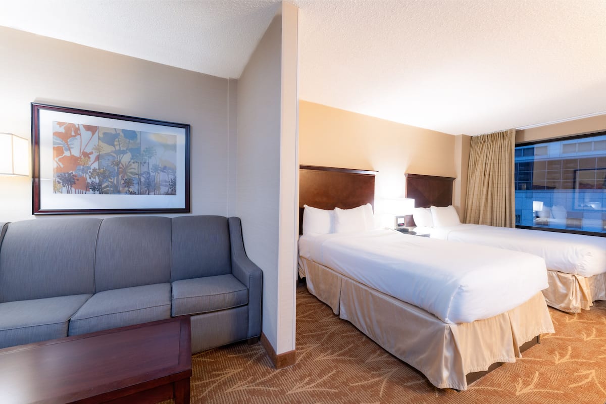 A hotel room with a sofa, two beds, a large window, and a framed artwork on the wall.