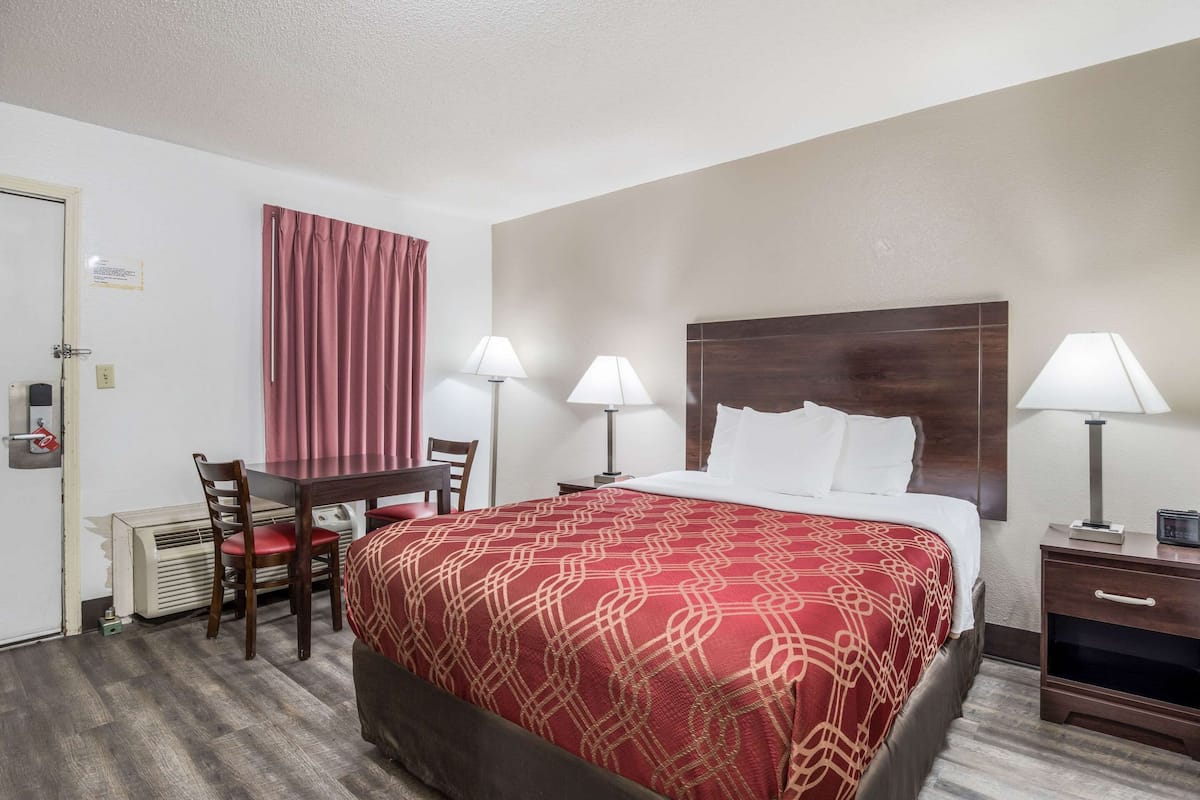 A hotel room with a large bed, two bedside tables with lamps, a desk with a chair, and a window with curtains.