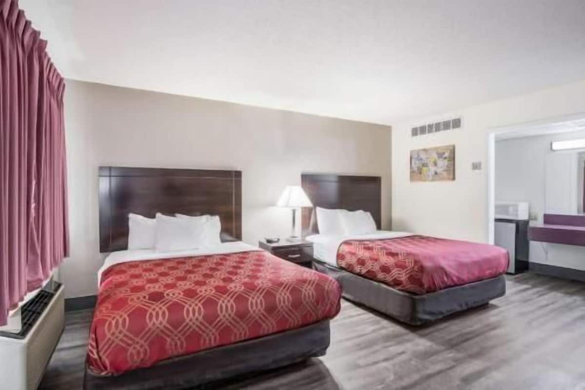 A hotel room with two beds, a nightstand with a lamp, a painting on the wall, and a bathroom visible through an open door.