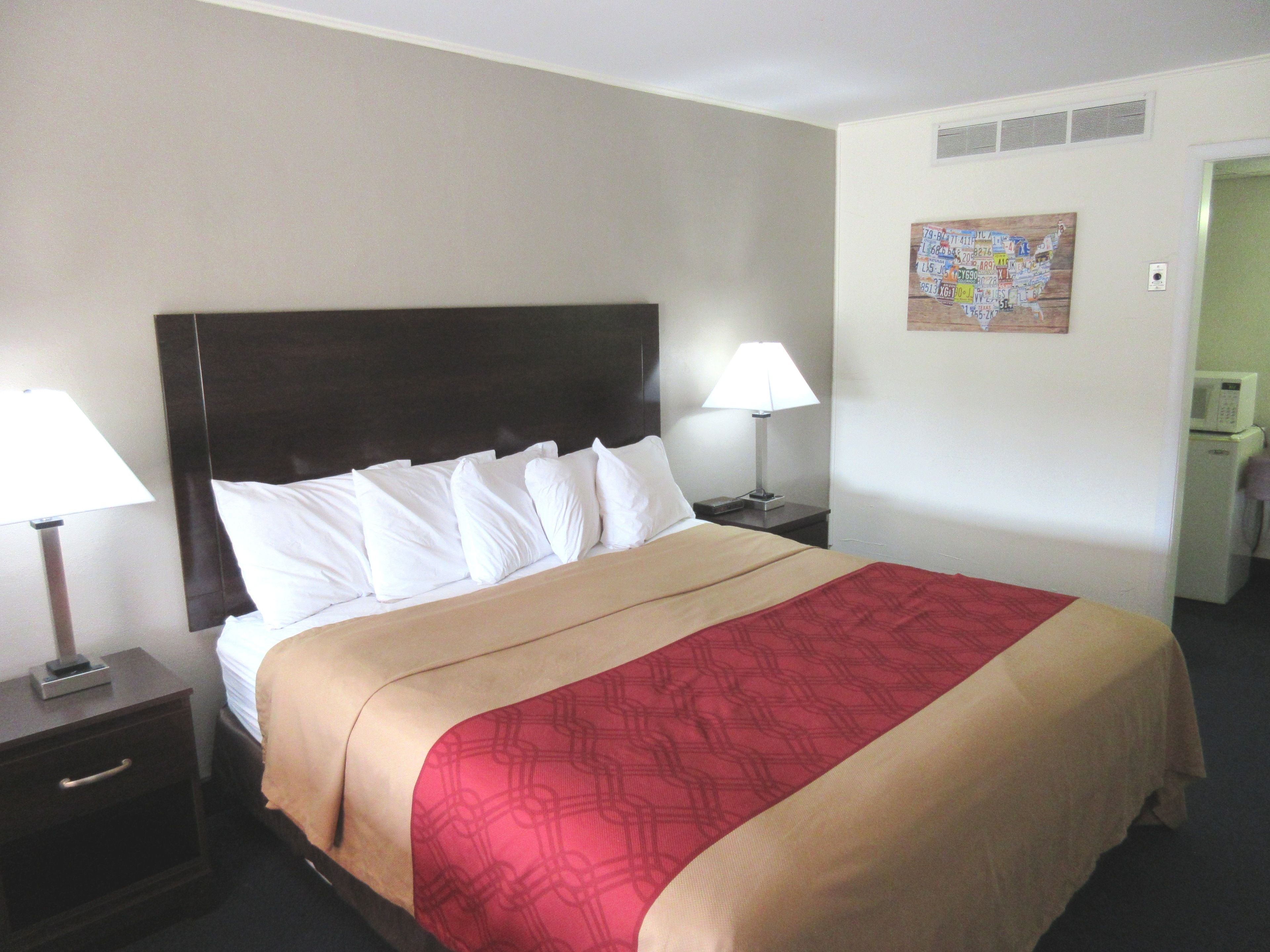 Una habitación de hotel con una cama grande, dos mesitas de noche con lámparas, un conducto de ventilación en la pared y un refrigerador pequeño.