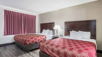 A hotel room with two beds, each with a red patterned bedspread, a wooden headboard, a bedside table with a lamp, and a window with red curtains.