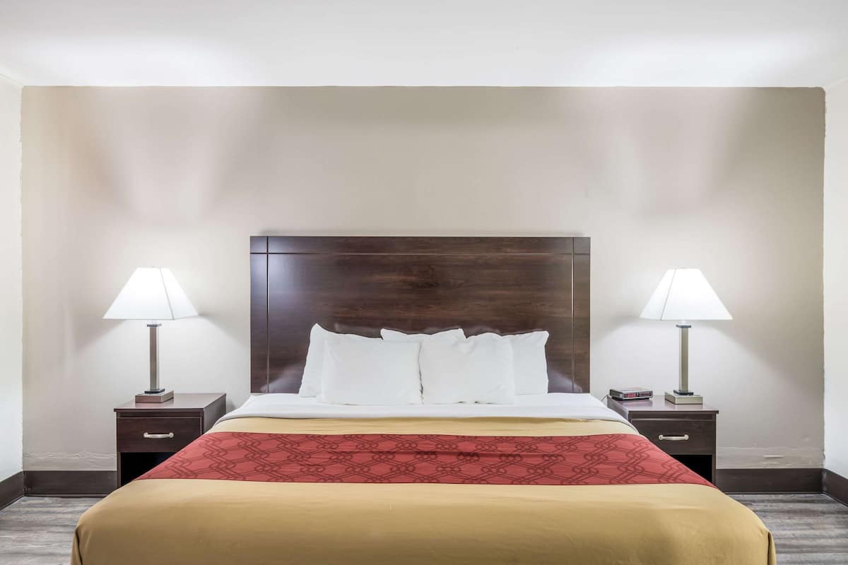 A hotel room with a large bed, two bedside tables with lamps, a wooden headboard, and a red patterned bedspread.