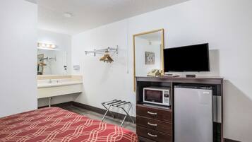 A hotel room with a bed, a dresser, a microwave, a refrigerator, a TV, a mirror, and a vanity with a sink.