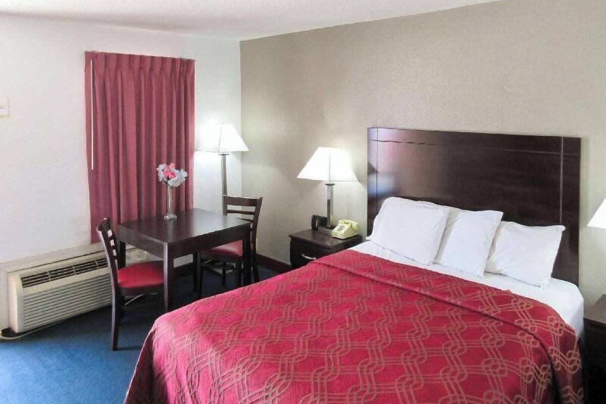 A hotel room with a bed, two bedside tables with lamps, a desk with a chair, a red curtain, and a wall-mounted heater.