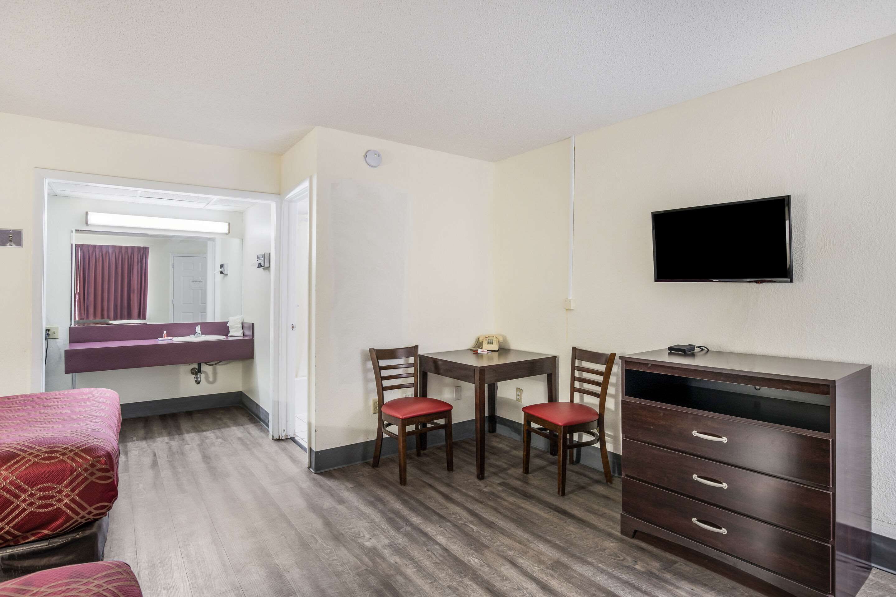 A hotel room with a bed, a desk with a chair, a dresser, a TV, and a bathroom visible through an open door.