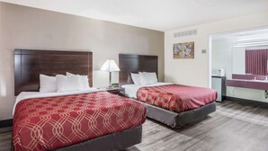 Habitación de hotel con dos camas, una mesita de noche con lámpara, una pequeña mesa, un espejo y una ventana.