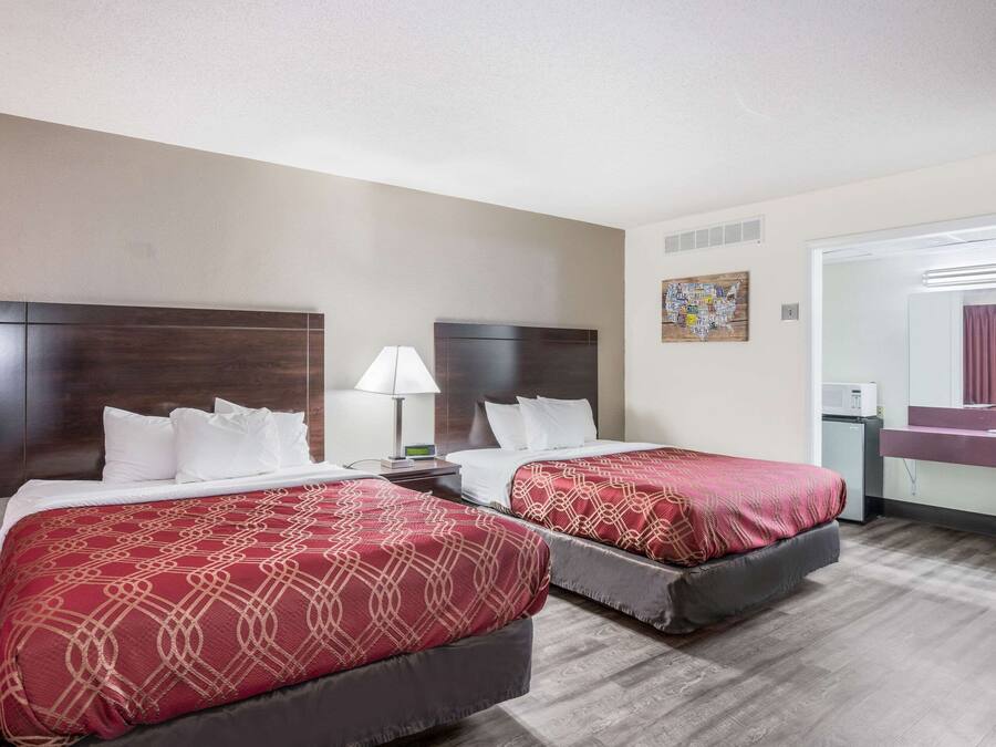 A hotel room with two beds, a nightstand with a lamp, a small table, a mirror, and a window.
