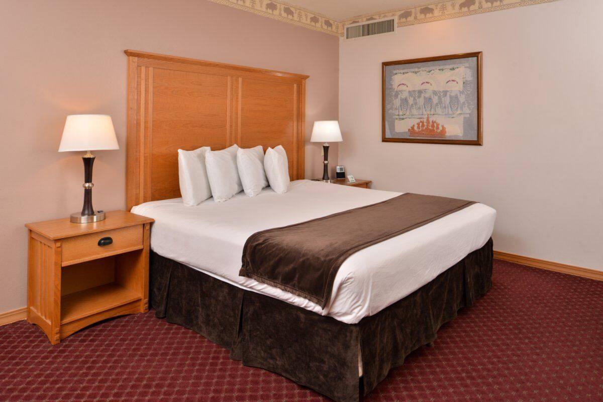 Quarto de hotel com uma cama grande, duas mesas de cabeceira com abajures, um cabeceira de madeira, um quadro na parede e um tapete estampado.
