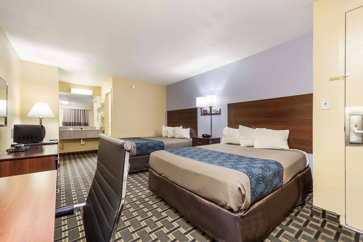 A hotel room with two beds, a desk, a chair, a TV, and a bathroom visible in the background.