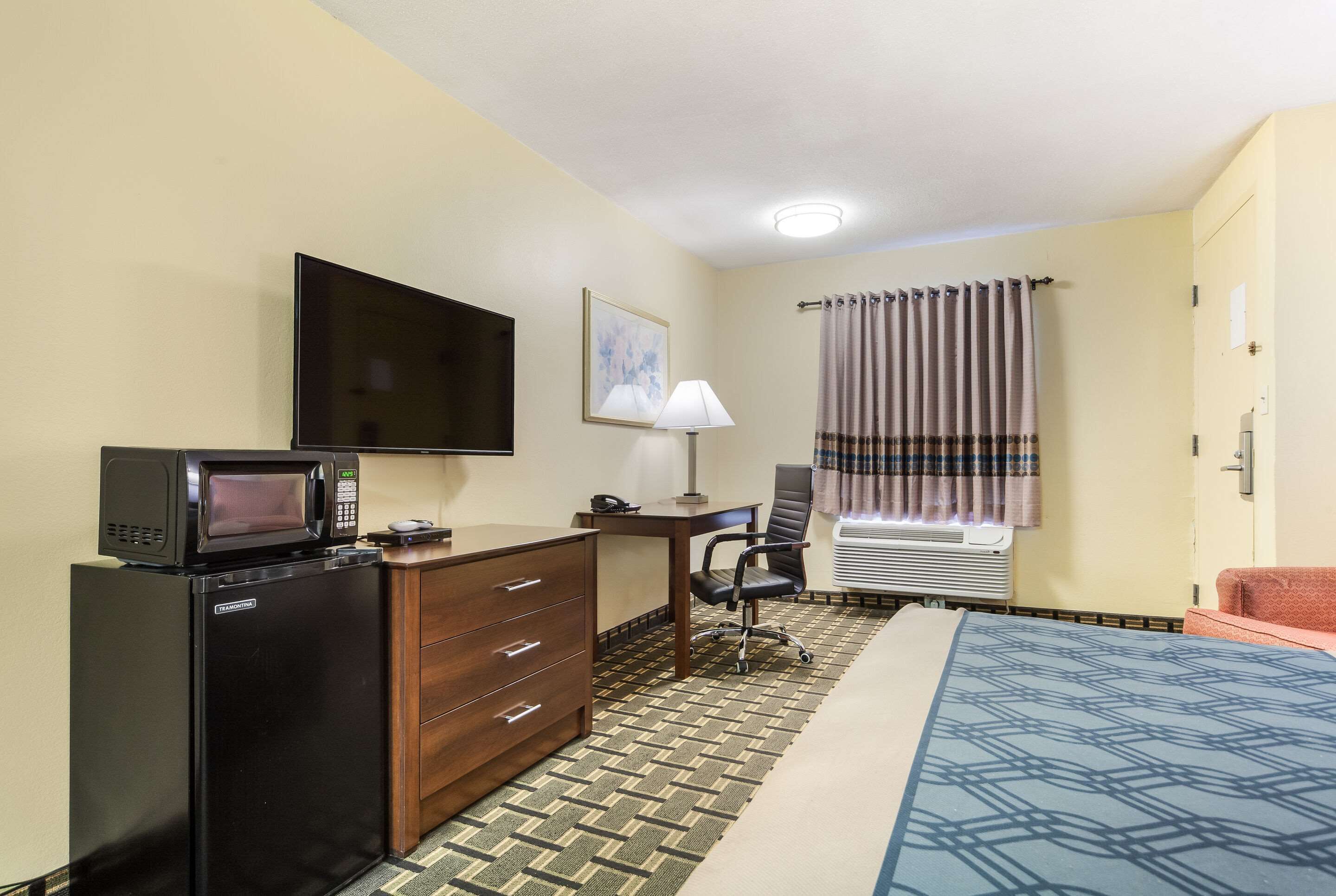 A hotel room with a bed, a desk with a chair, a television, a microwave, a dresser, a window with curtains, and an air conditioning unit.