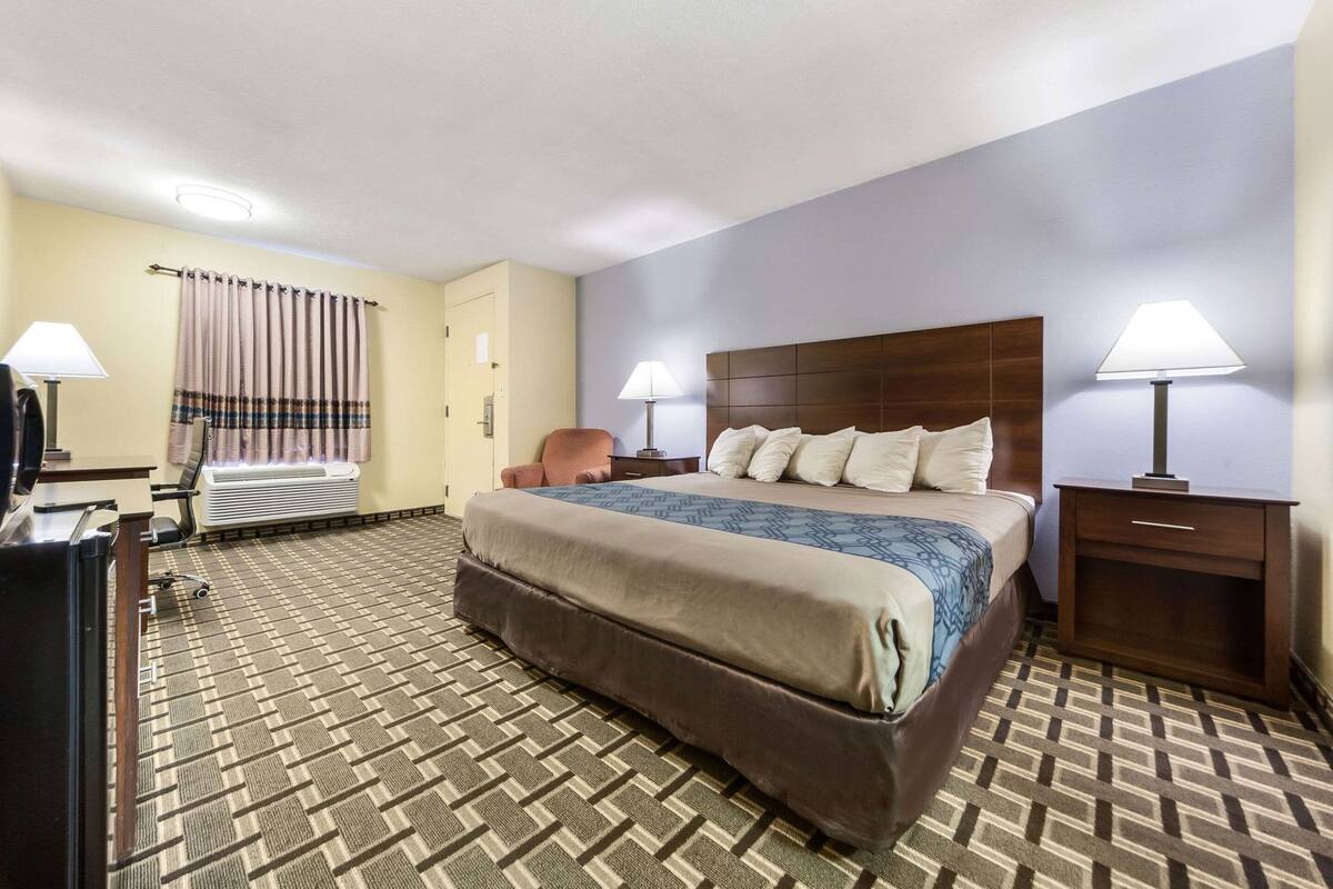 A hotel room with a large bed, two bedside tables with lamps, a desk with a chair, an air conditioning unit, and a patterned carpet.