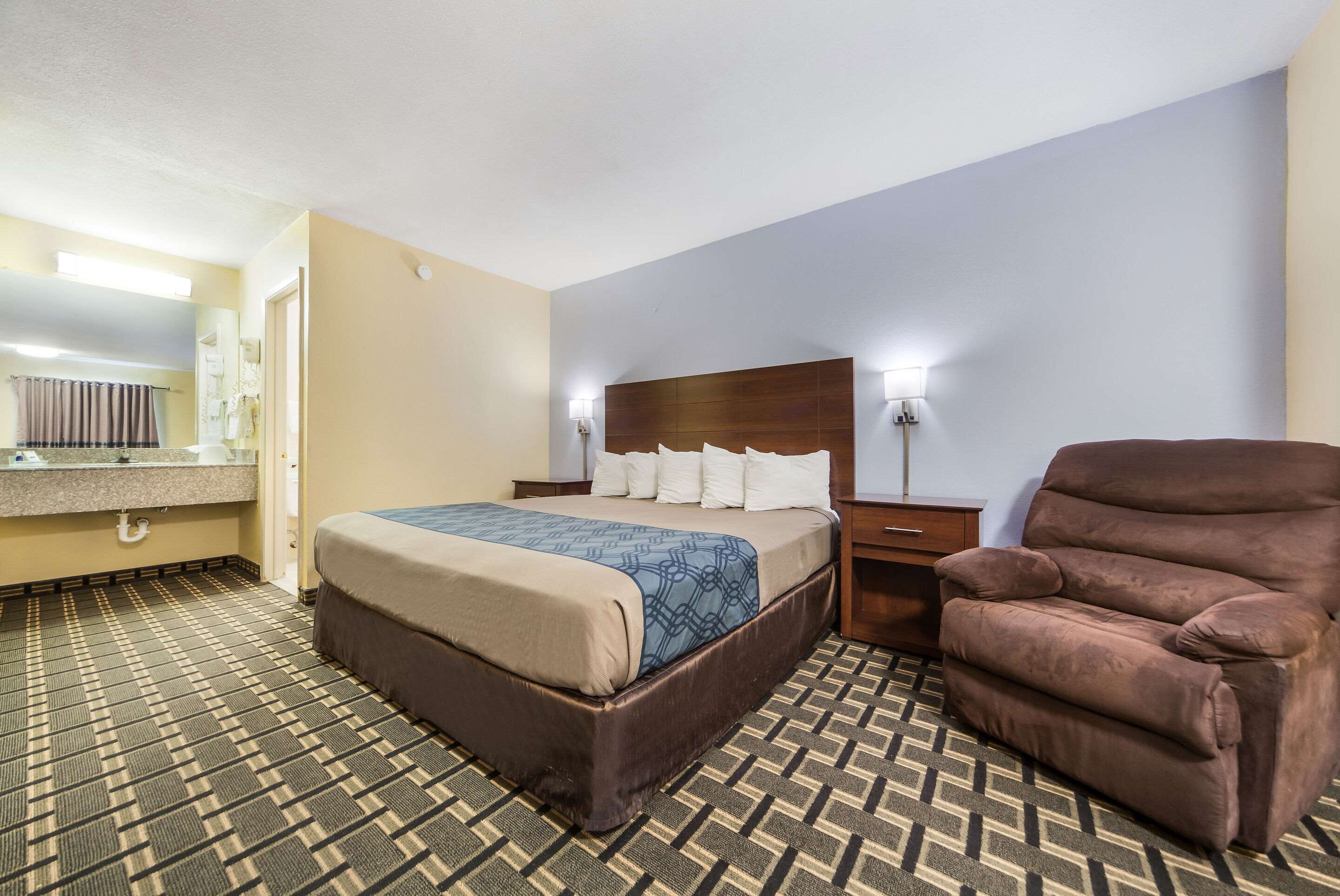 Habitación de hotel con una cama, un sillón marrón, una mesita de noche y un baño con espejo y perchero.
