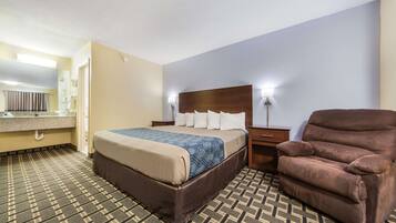 Habitación de hotel con una cama, un sillón marrón, una mesita de noche y un baño con espejo y perchero.