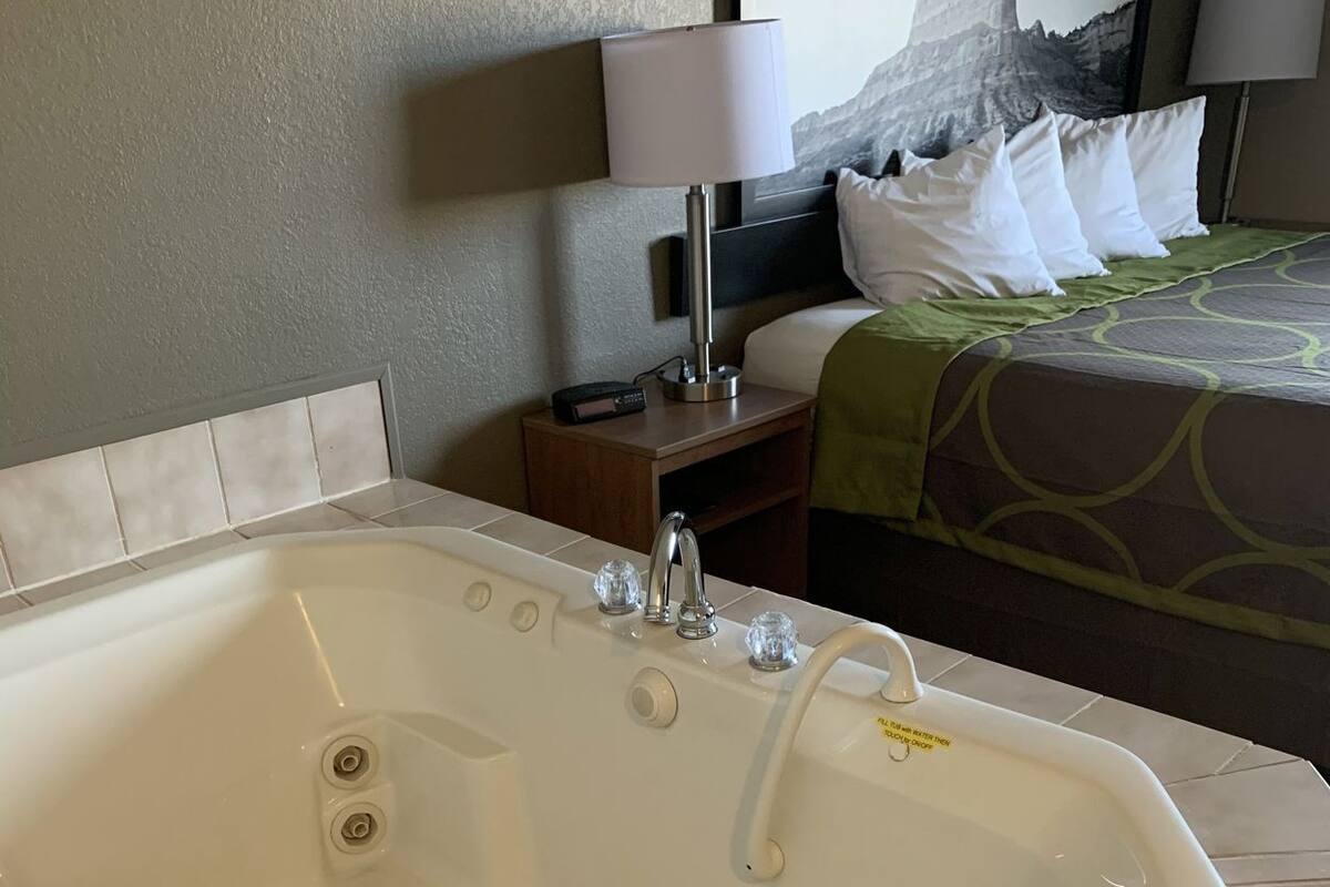 Une chambre d’hôtel avec une grande baignoire, un lit recouvert d’une couvre-lit à motifs verts et blancs, une table de chevet avec une lampe et un tableau accroché au mur.