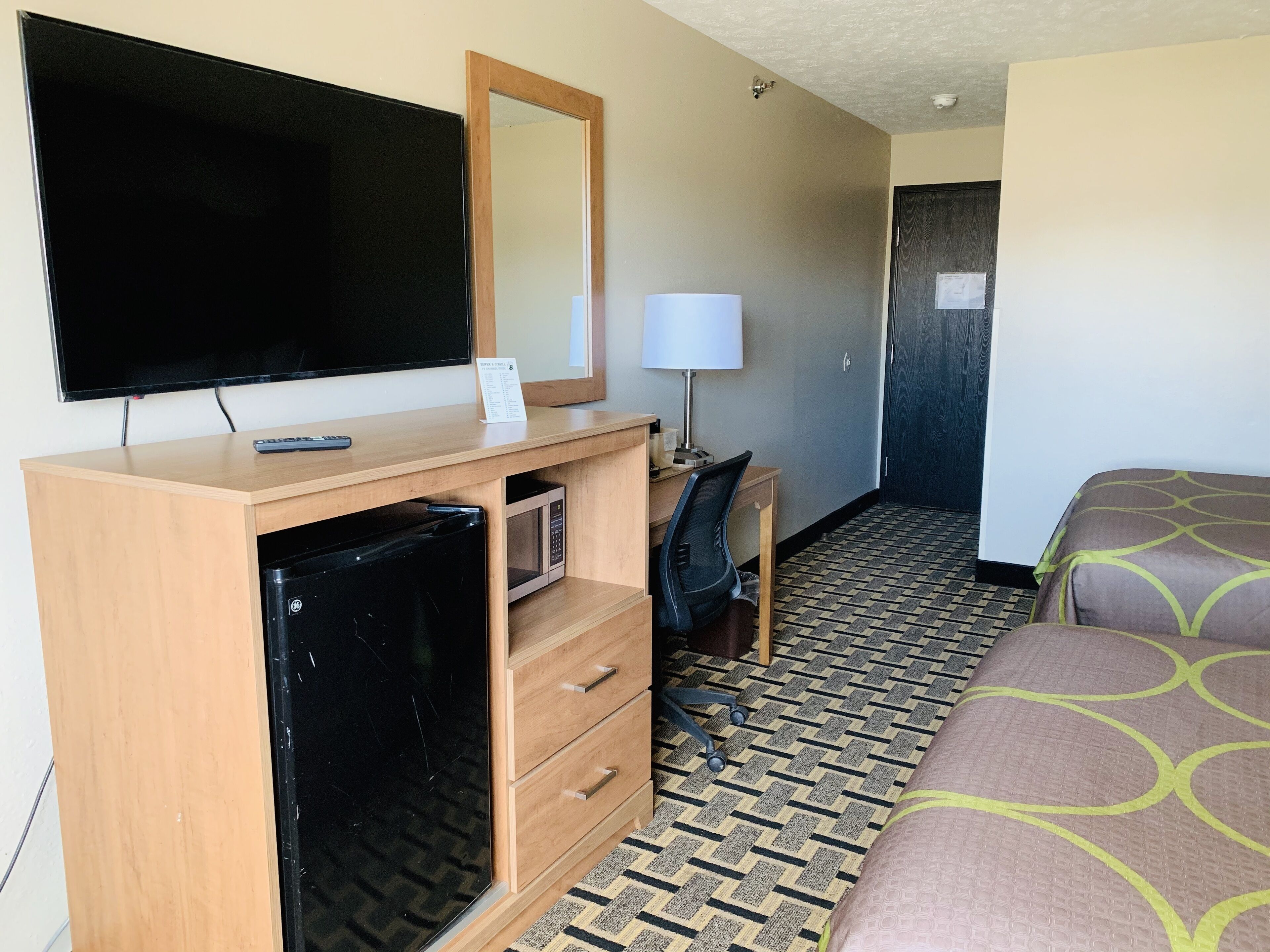 A hotel room with a flat-screen TV, a wooden desk with a lamp, a microwave, a mini fridge, and a bed with patterned bedding.