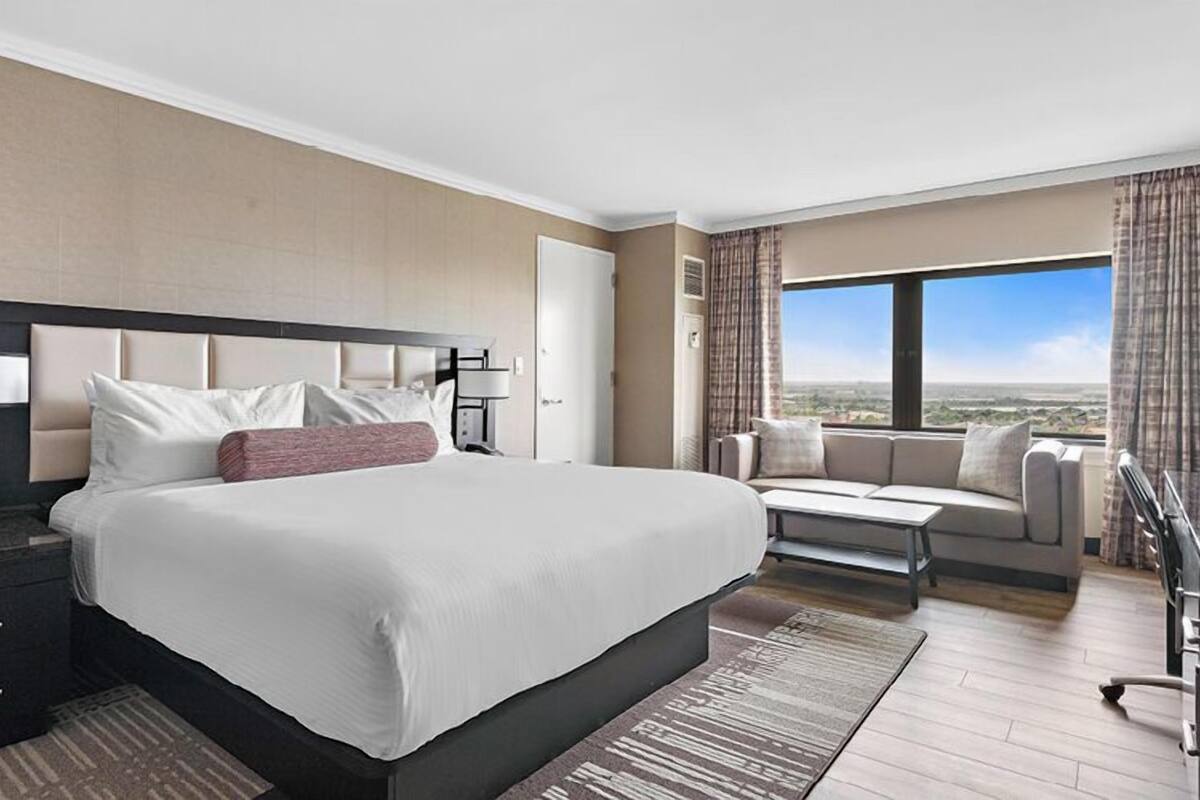 A hotel room with a large bed, a desk with a lamp, a sofa, and a view of the city.