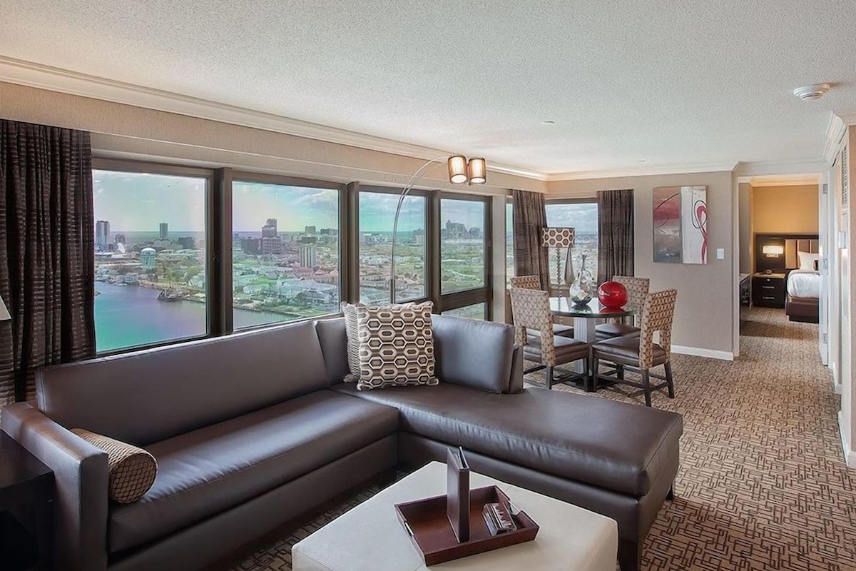 A hotel room with a large window, a sofa, a dining area, and a bed.