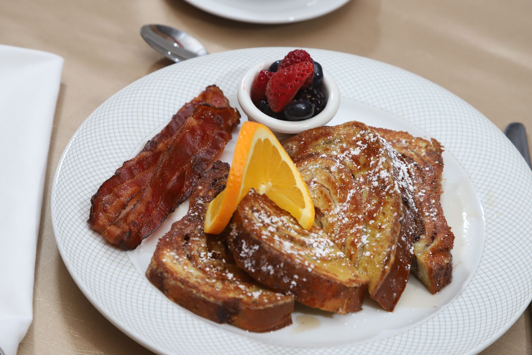 A plate of French toast with powdered sugar, a slice of orange, and a small bowl of berries.