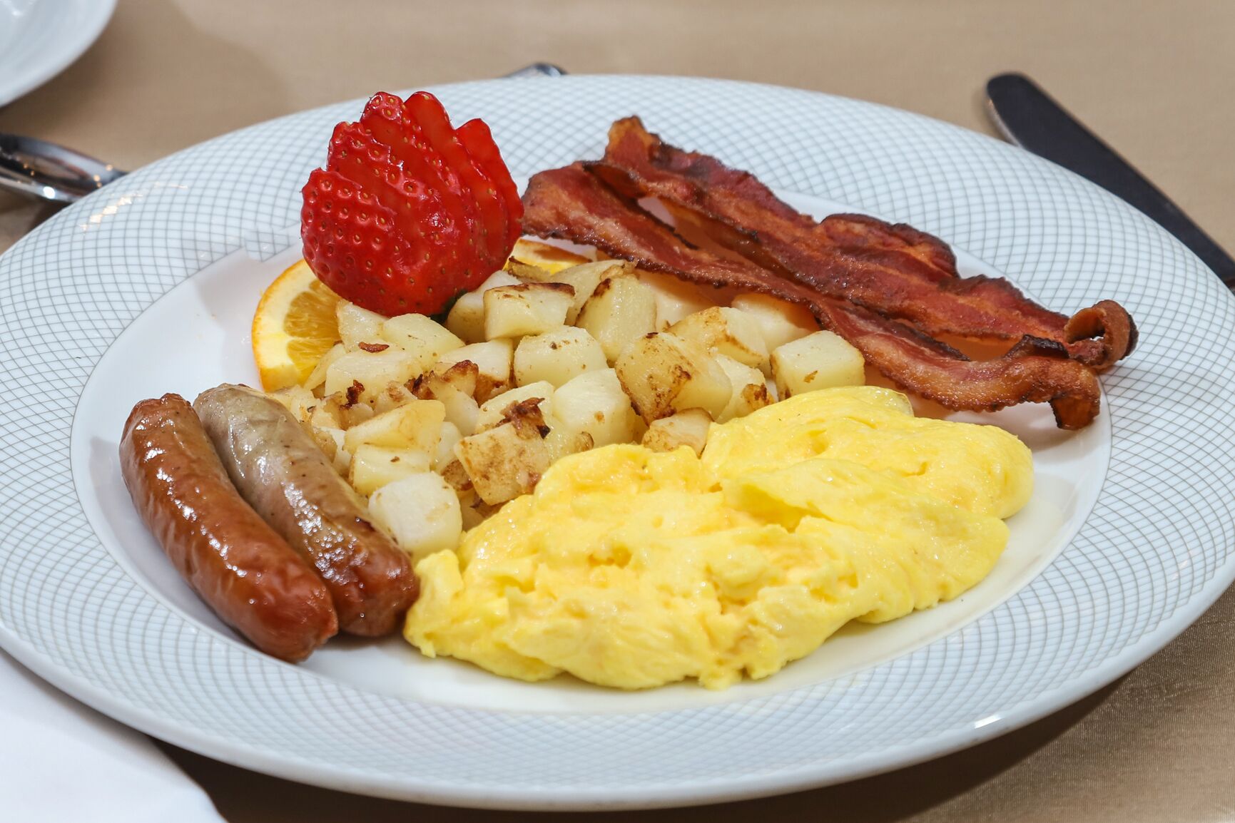 Un plato con huevos revueltos, tocino, salchichas, patatas asadas y una fresa.