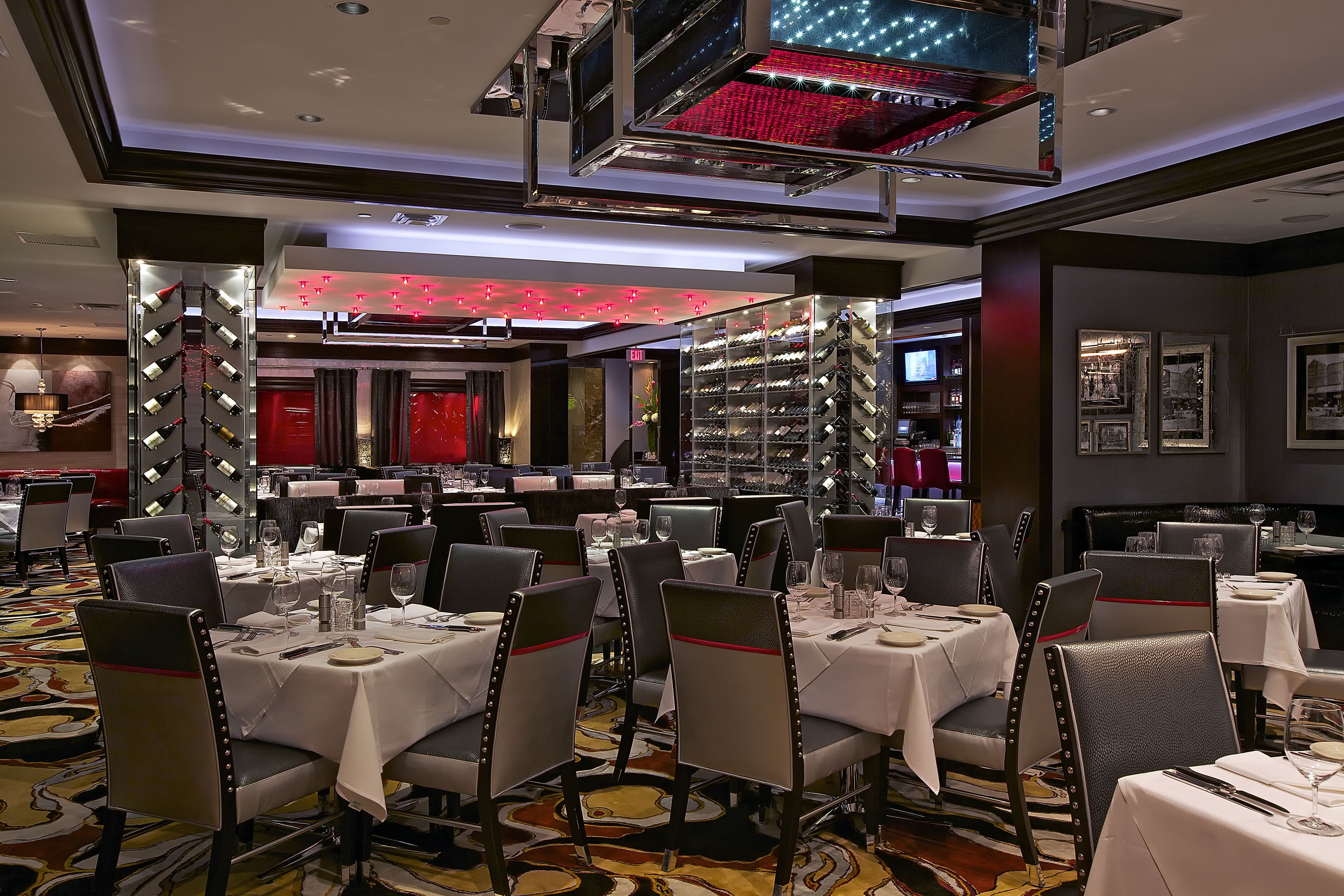 A hotel dining area with multiple tables and chairs, a wine rack, and a red-lit wall.