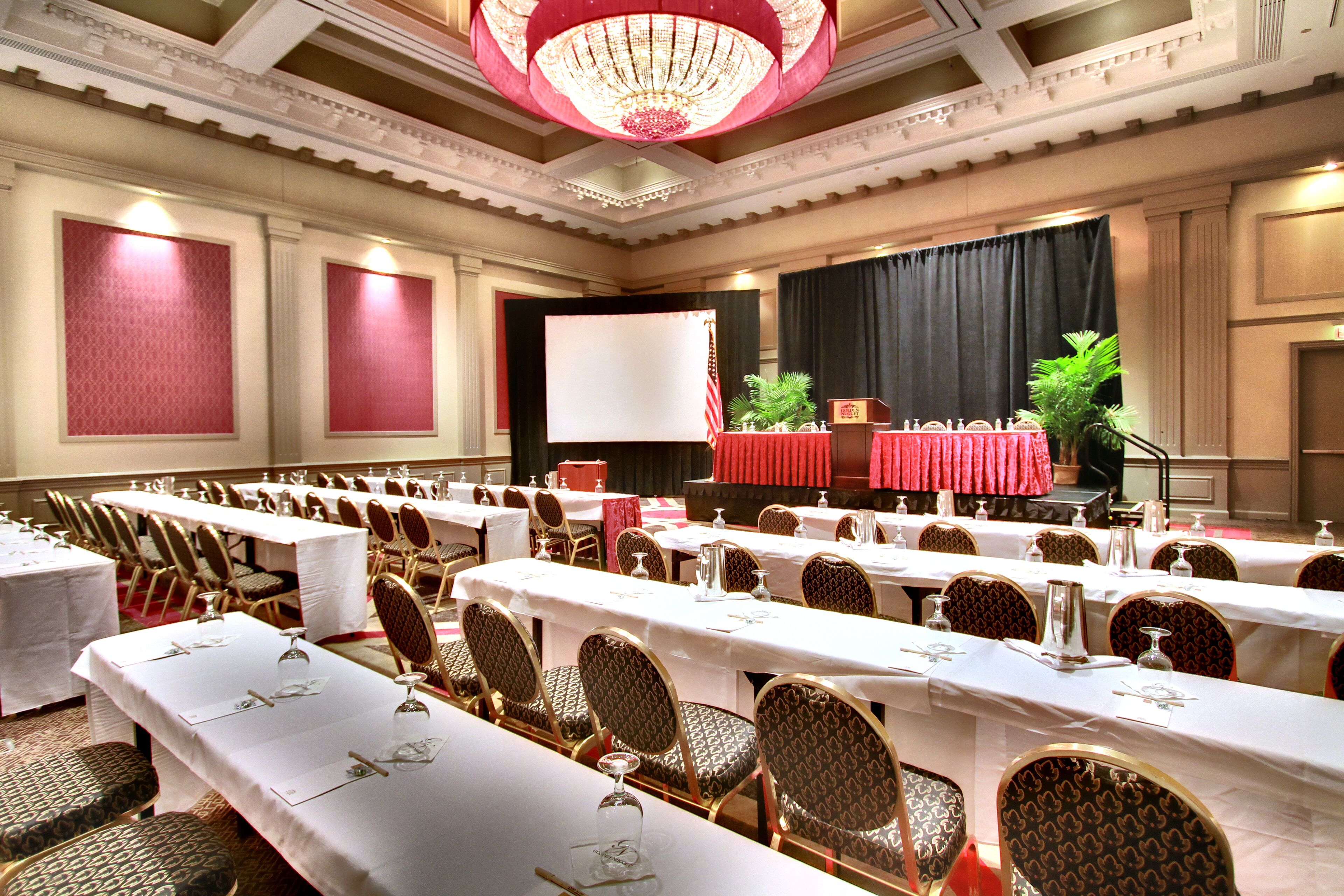 A banquet hall with long tables, chairs, a stage, and a large screen.