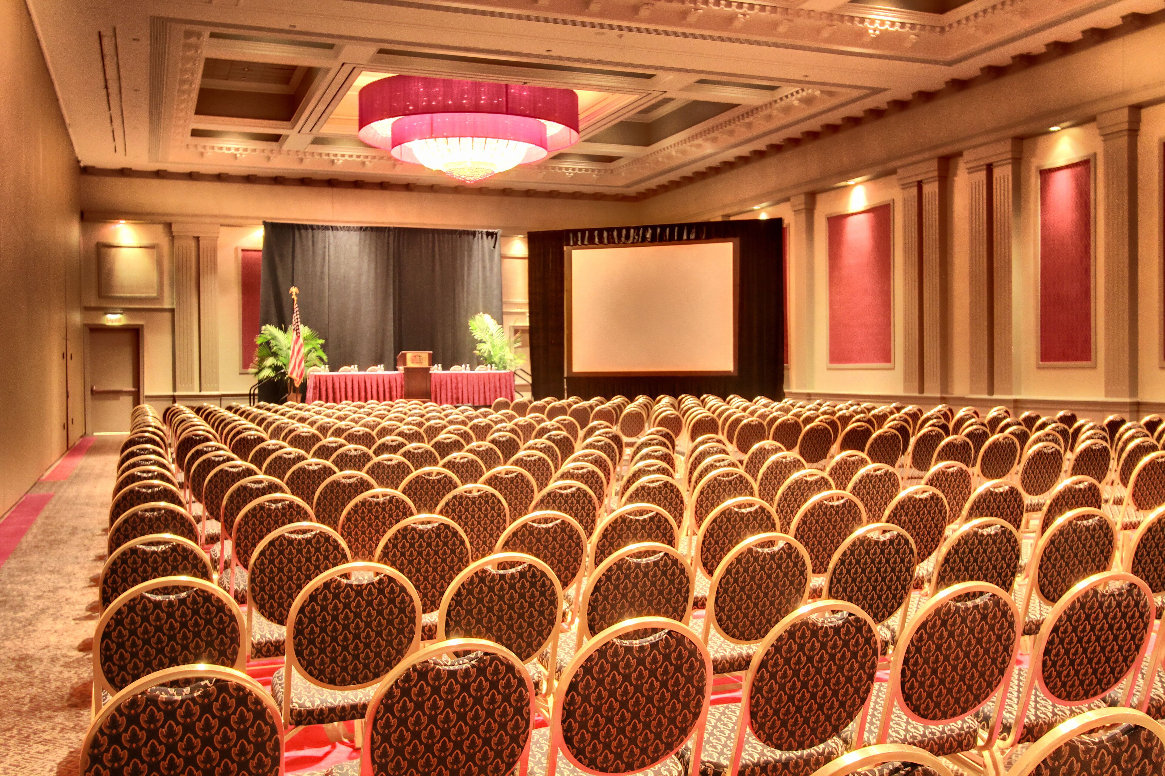 A conference room with rows of patterned chairs, a stage with a red table, and a large screen.