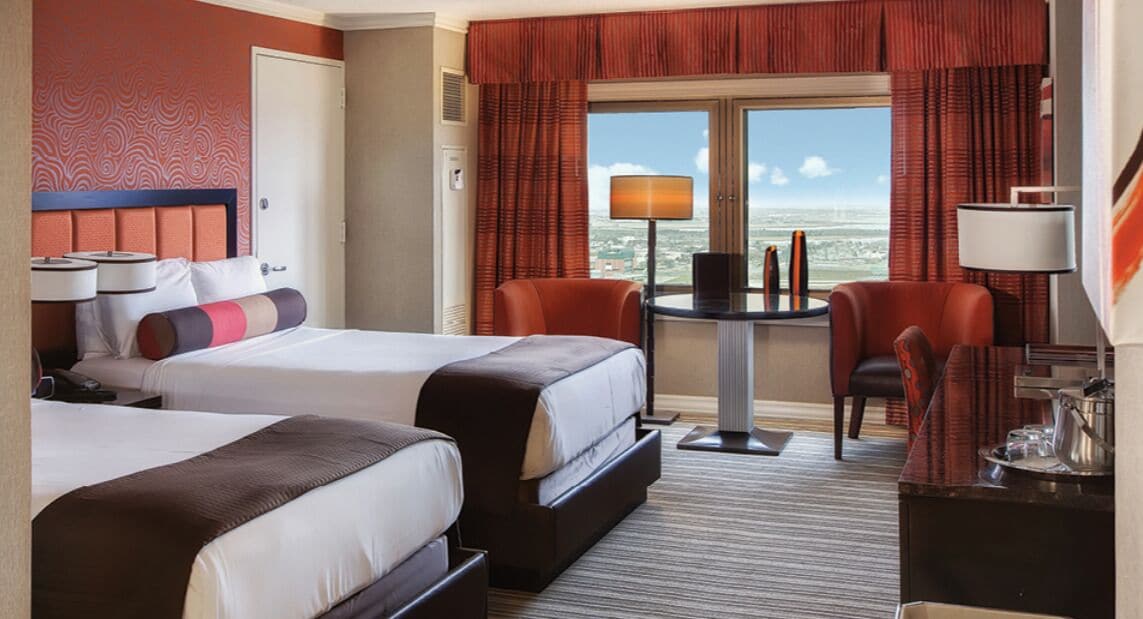 A hotel room with two beds, a small table with a lamp, a chair, and a view of the city through large windows.