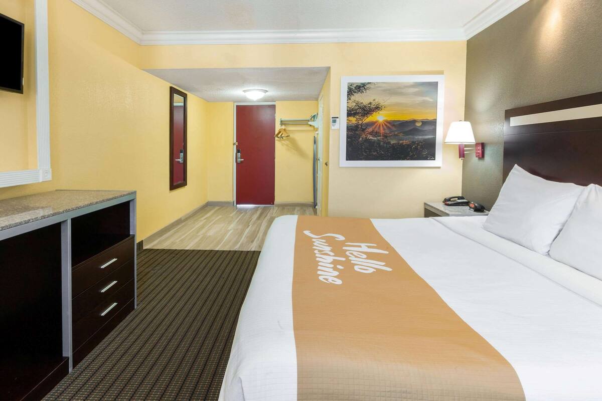 A hotel room with a bed, a desk, a mirror, a door, and a painting on the wall.