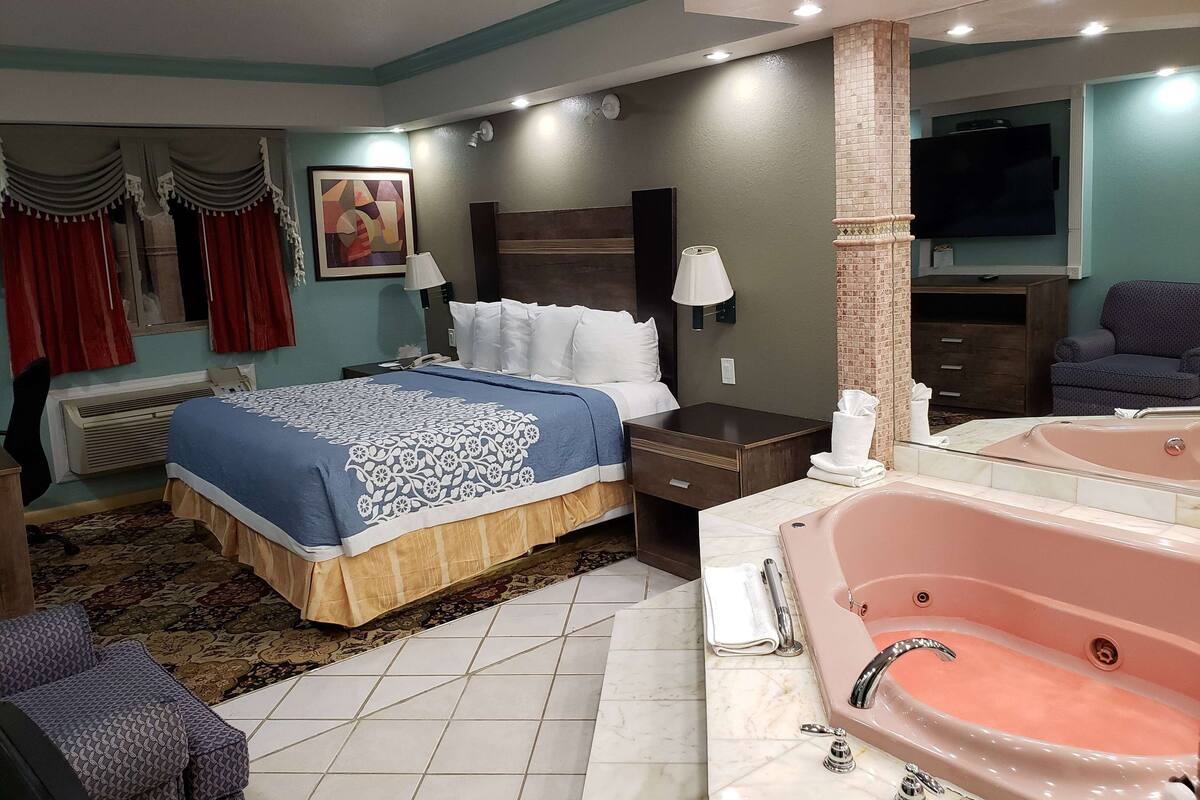 A hotel room with a large bed, a sitting area with a chair, a bathroom with a bathtub, and a television on the wall.