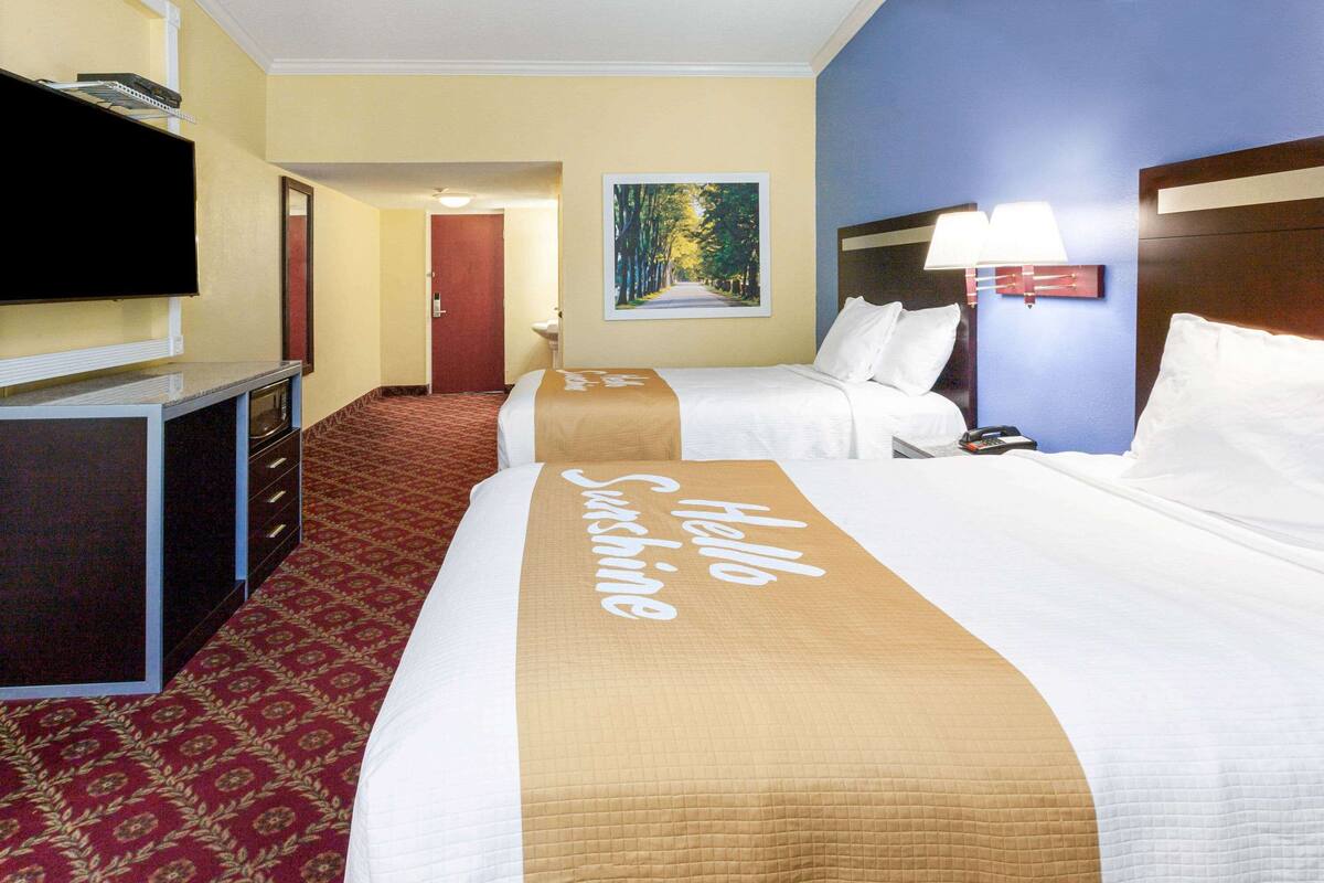A hotel room with two beds, a television, a microwave, a painting of a forest, and a 'Hello Sunshine' bed runner.