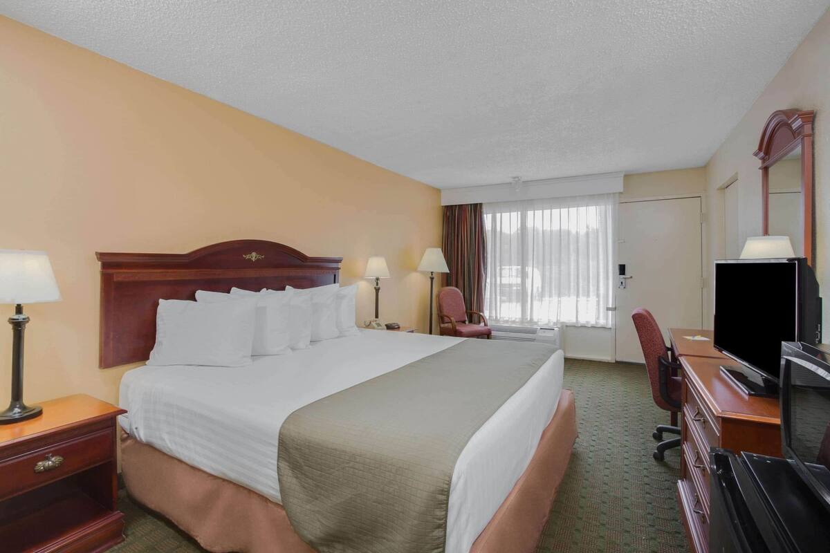 A hotel room with a large bed, a desk with a television, a chair, and a lamp.