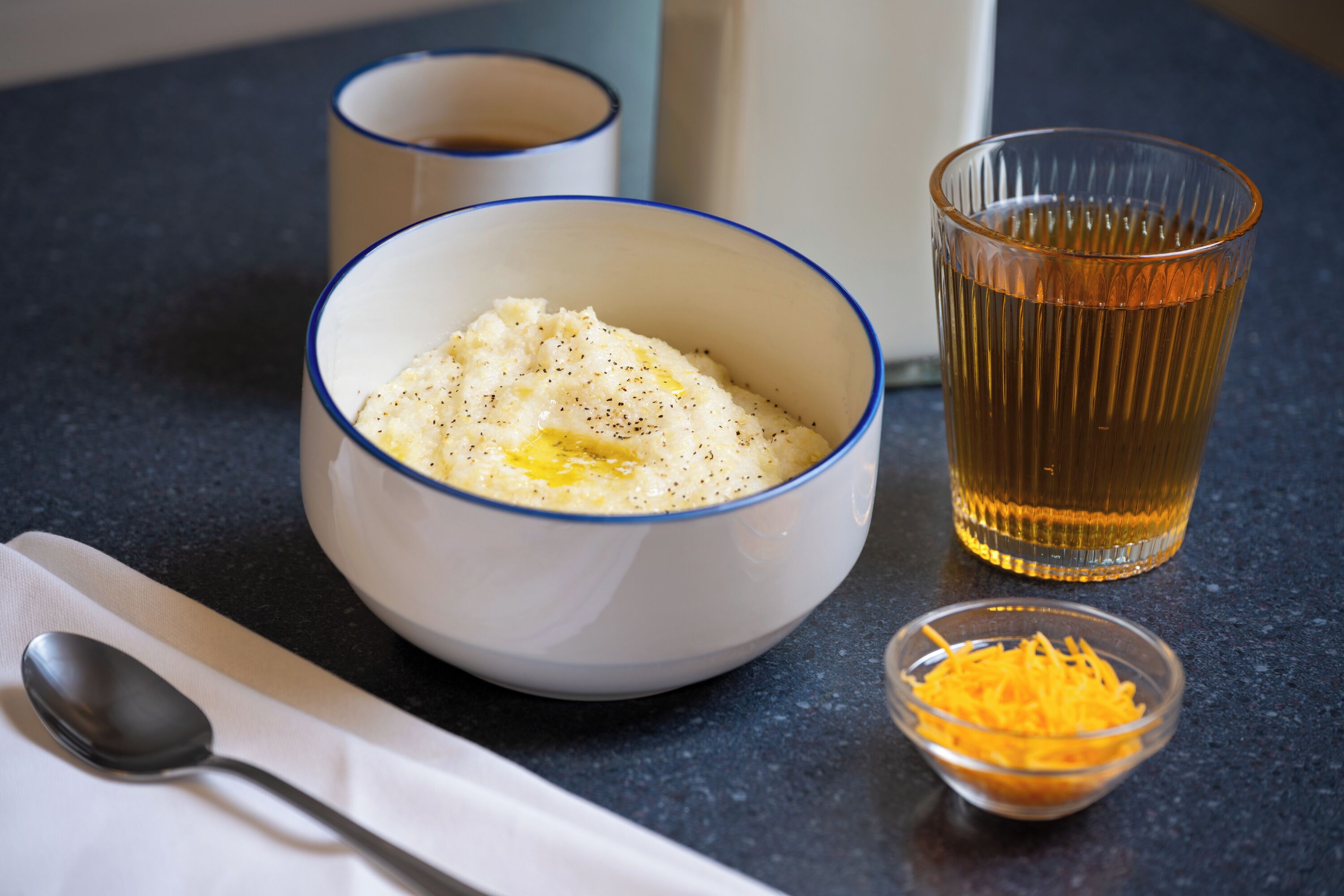 A bowl of mashed potatoes with a spoon and a small bowl of shredded cheese on a dark countertop.