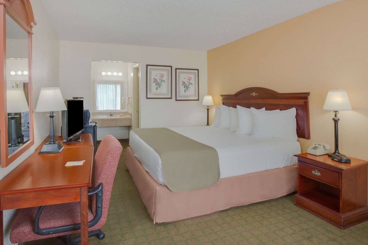 A hotel room with a bed, desk, chair, bedside table, lamp, mirror, and framed pictures on the wall.