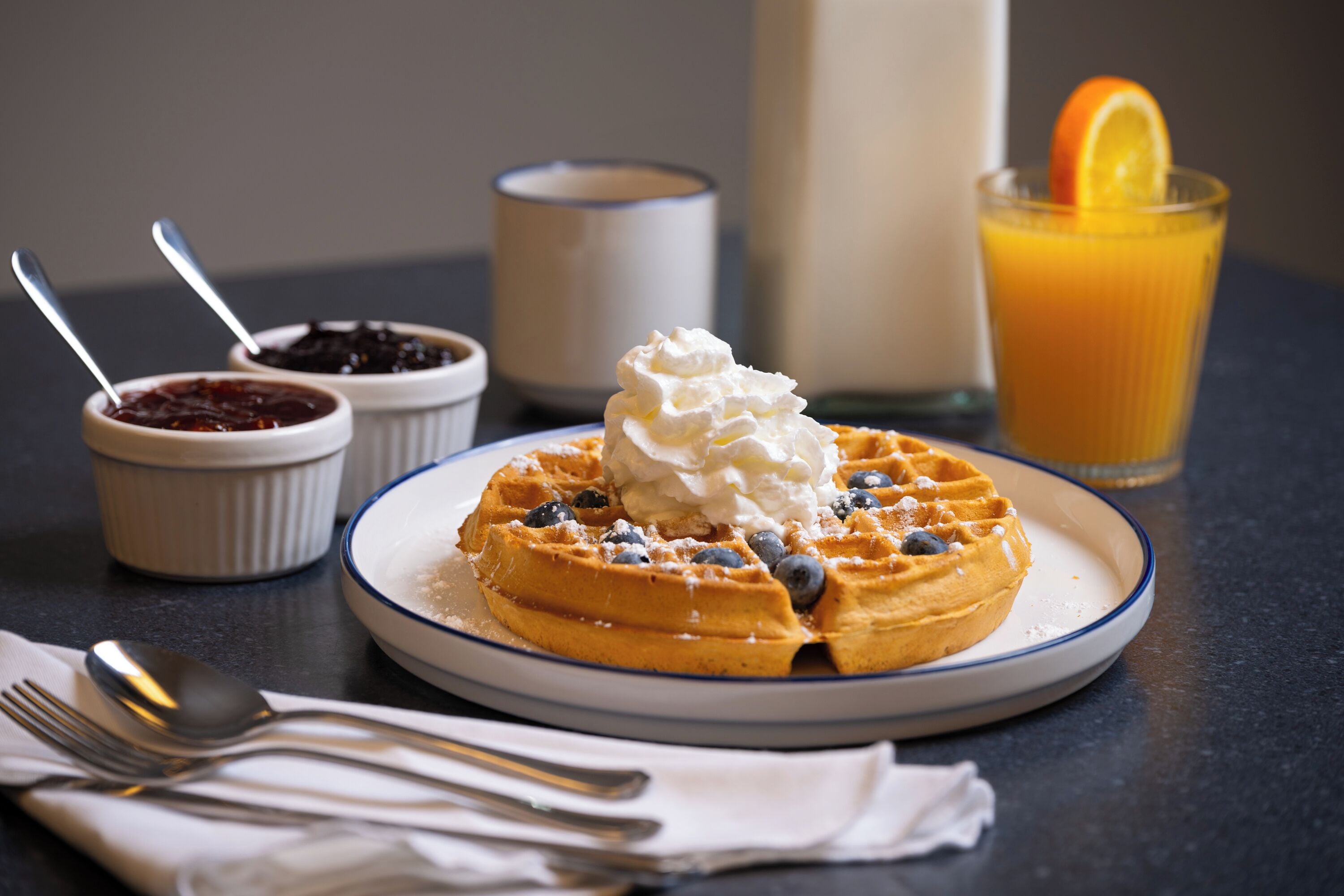 A breakfast plate with waffles, whipped cream, blueberries, and a glass of orange juice.