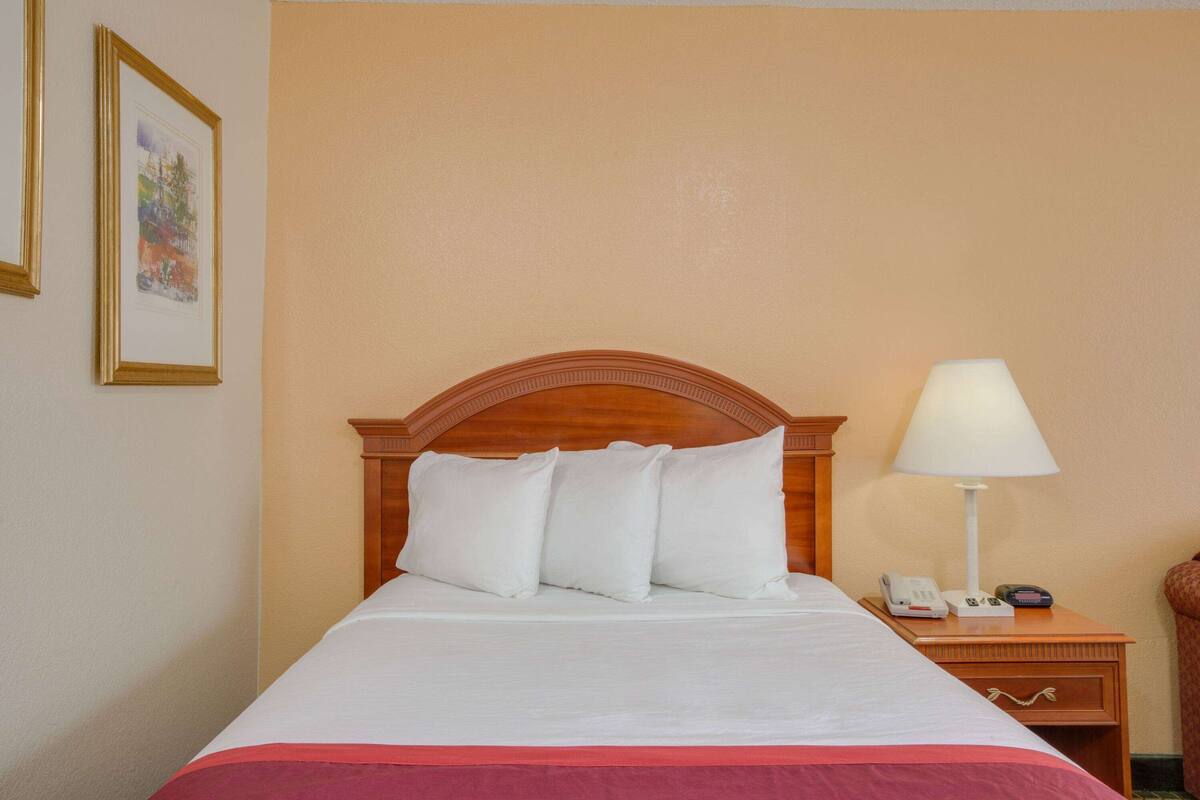 A neatly made bed with white bedding and a red accent at the foot. A wooden headboard with a carved design tops the bed. A bedside table with a lamp and a telephone is visible to the right.