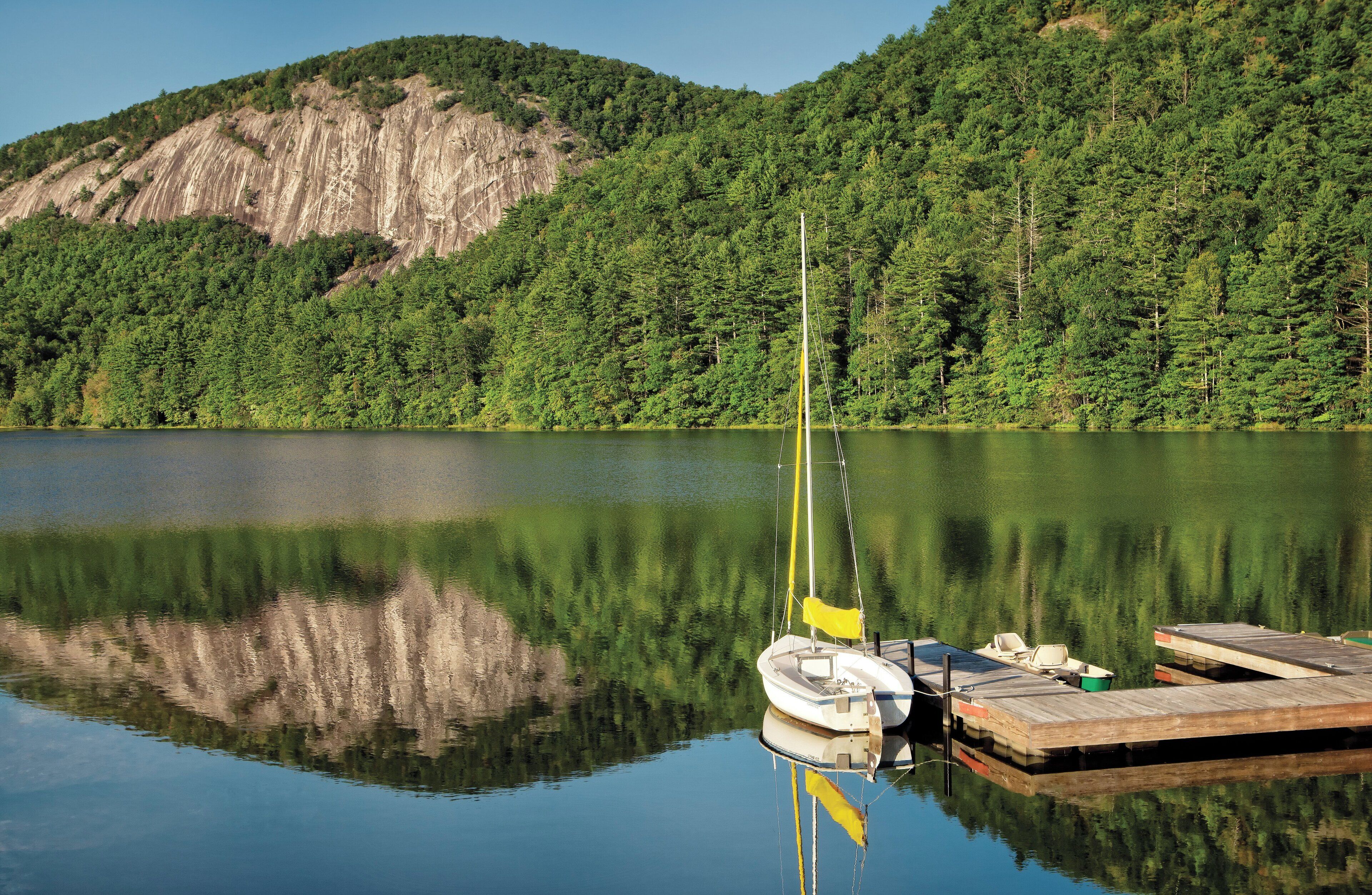 A calm lake with a dock and a sailboat, surrounded by dense forest and a rocky hill.