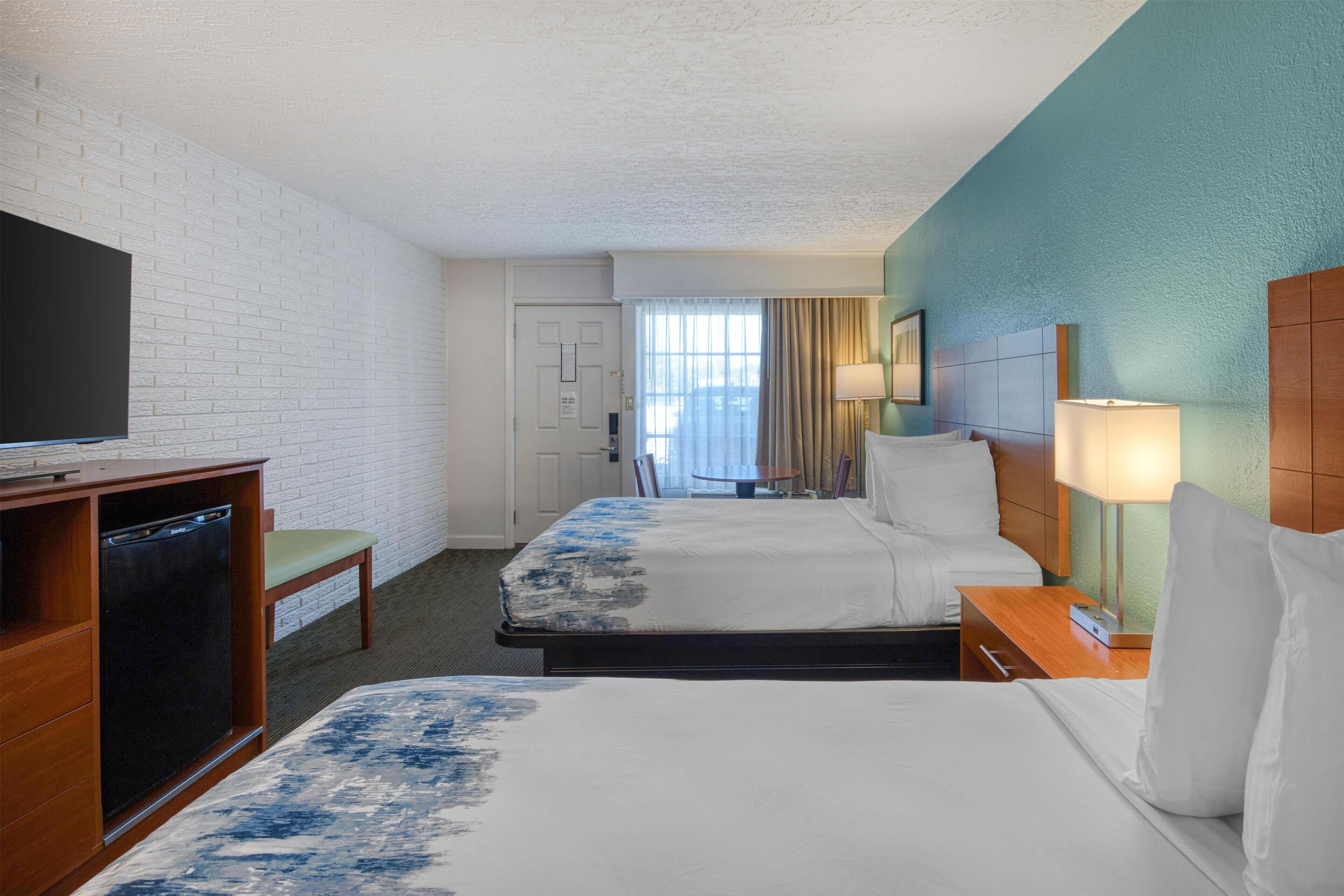 Quarto de hotel com duas camas, uma televisão, uma escrivaninha com abajur e vista para o exterior através de uma janela.