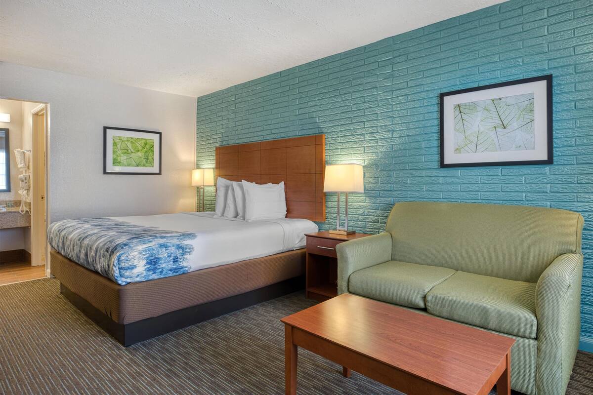 A hotel room with a bed, a green armchair, a wooden table, and two framed pictures on the wall.