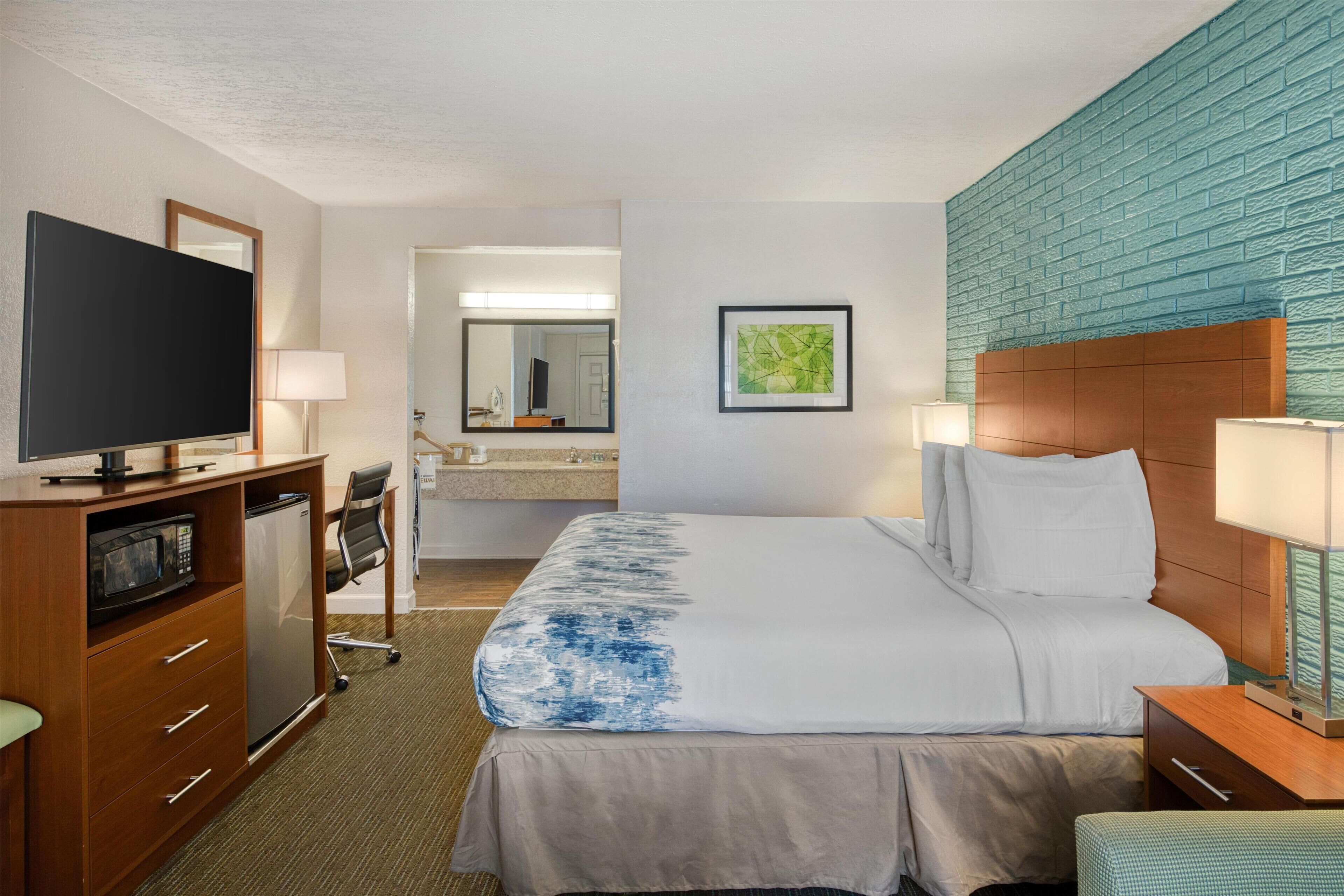 Ein Hotelzimmer mit Bett, Fernseher, Schreibtisch, Stuhl, Mikrowelle, Kühlschrank und einem Badezimmer mit Spiegel, sowie einer blauen Ziegelwand.