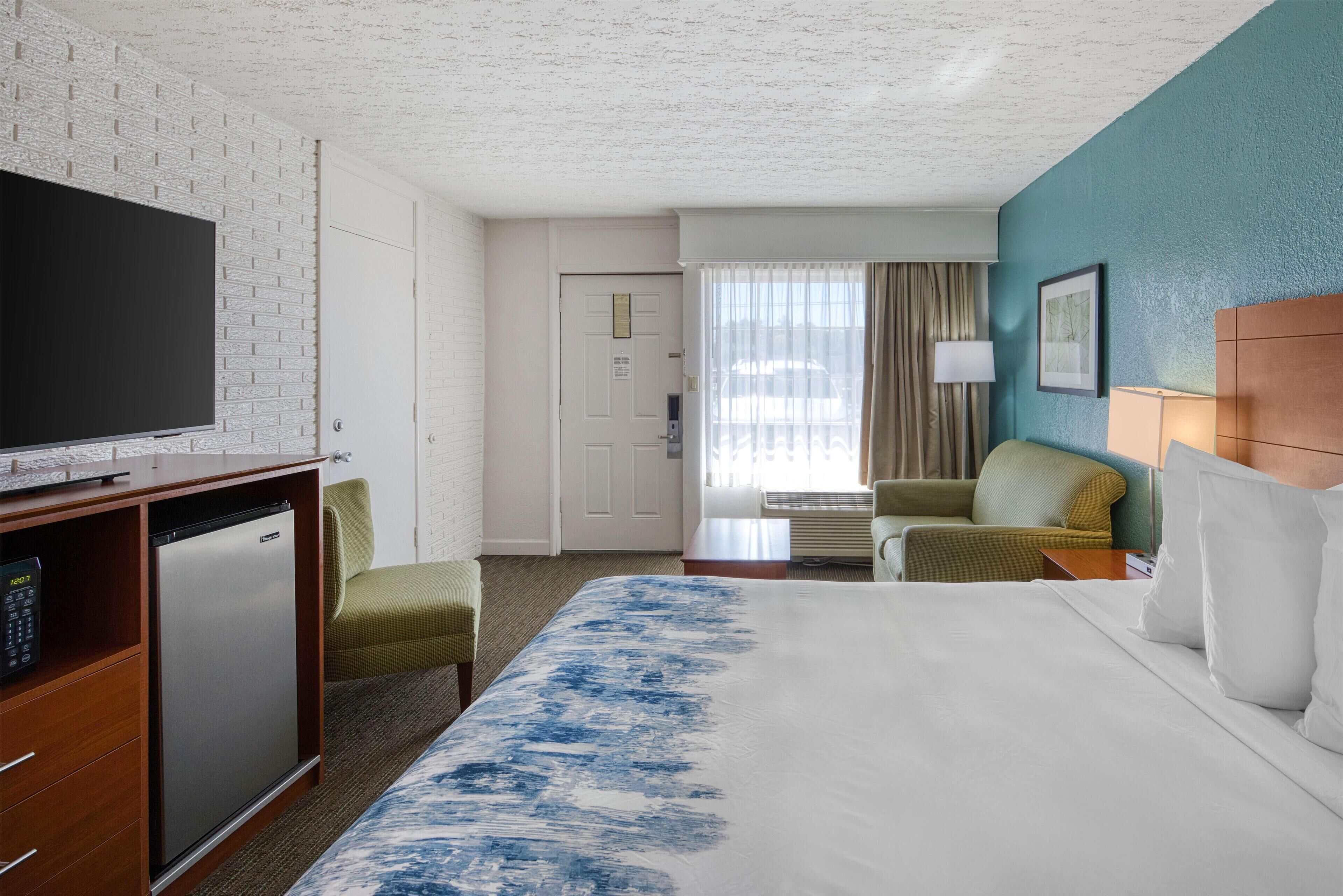 A hotel room with a large bed, a TV, a mini-fridge, a desk, and a seating area with a chair and a table.
