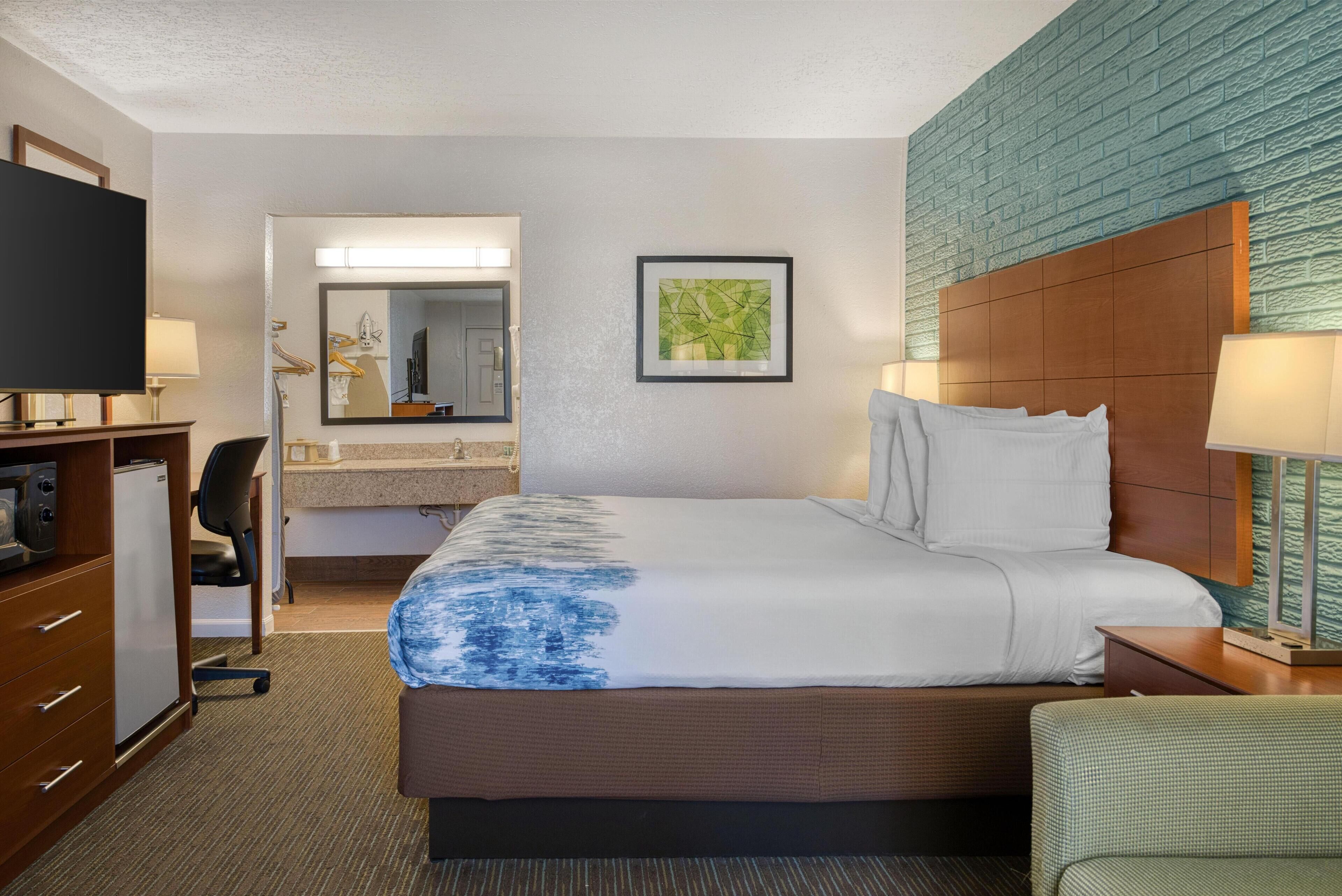 A hotel room with a large bed, a desk with a computer, a TV, and a bathroom with a mirror and sink.