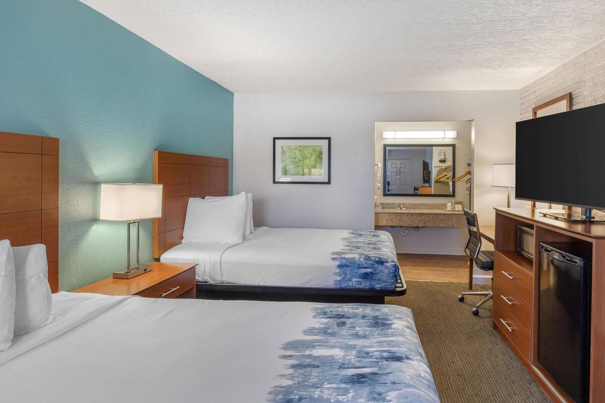 A hotel room with two beds, a desk, a television, a mirror, and a bathroom visible through an open door.