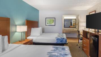 Une chambre d’hôtel avec deux lits, un bureau, une télévision, un miroir et une salle de bain visible à travers une porte ouverte.