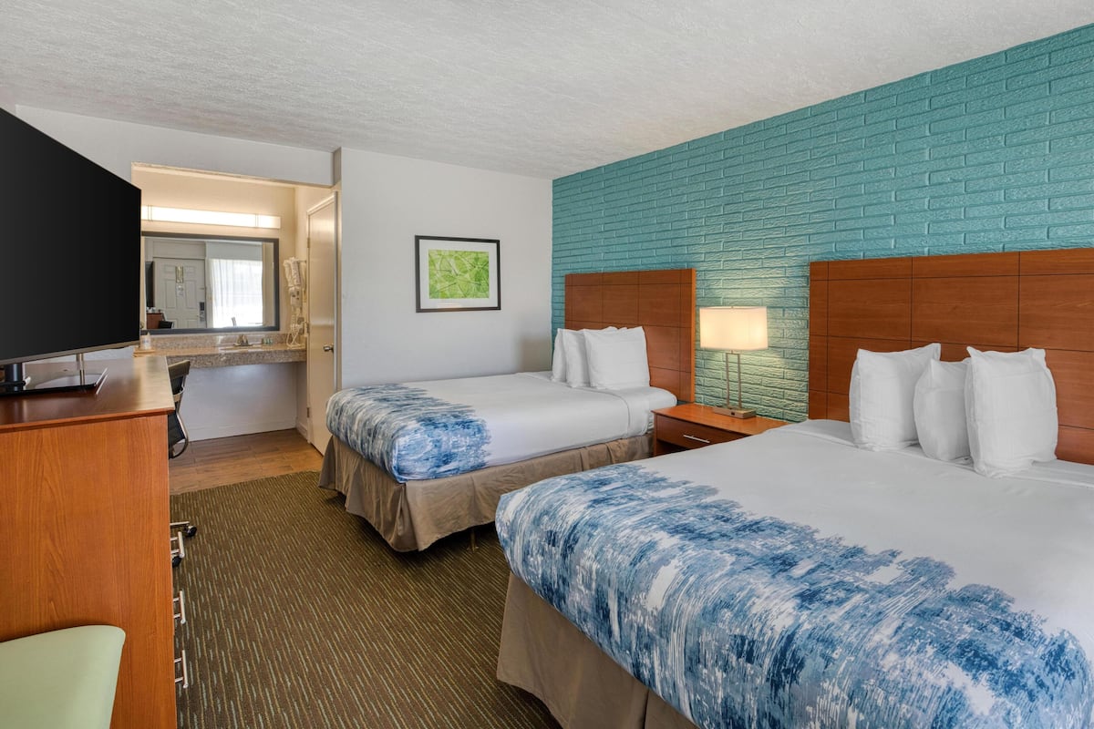 A hotel room with two beds, a desk, a TV, a bathroom, and a teal brick wall.