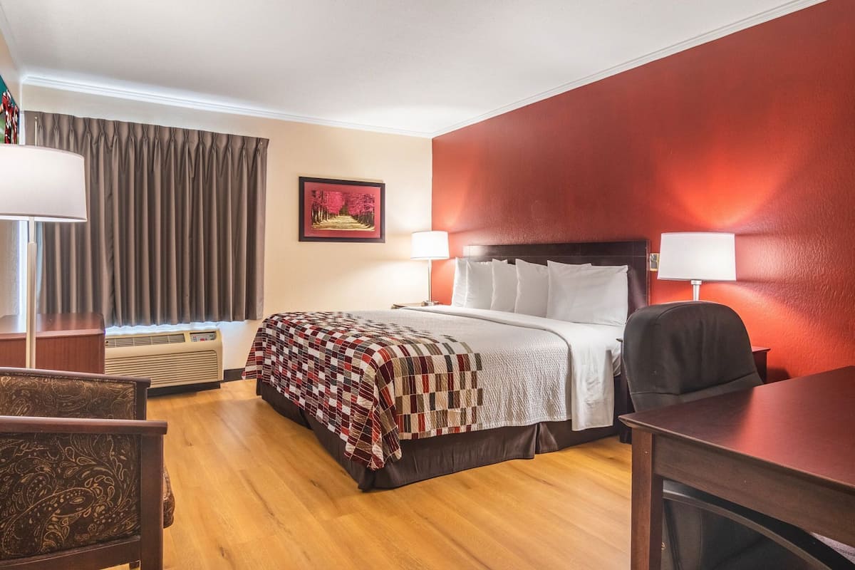 A hotel room with a bed, bedside lamps, a desk, a chair, a window with curtains, an air conditioner, and a wall-mounted picture.