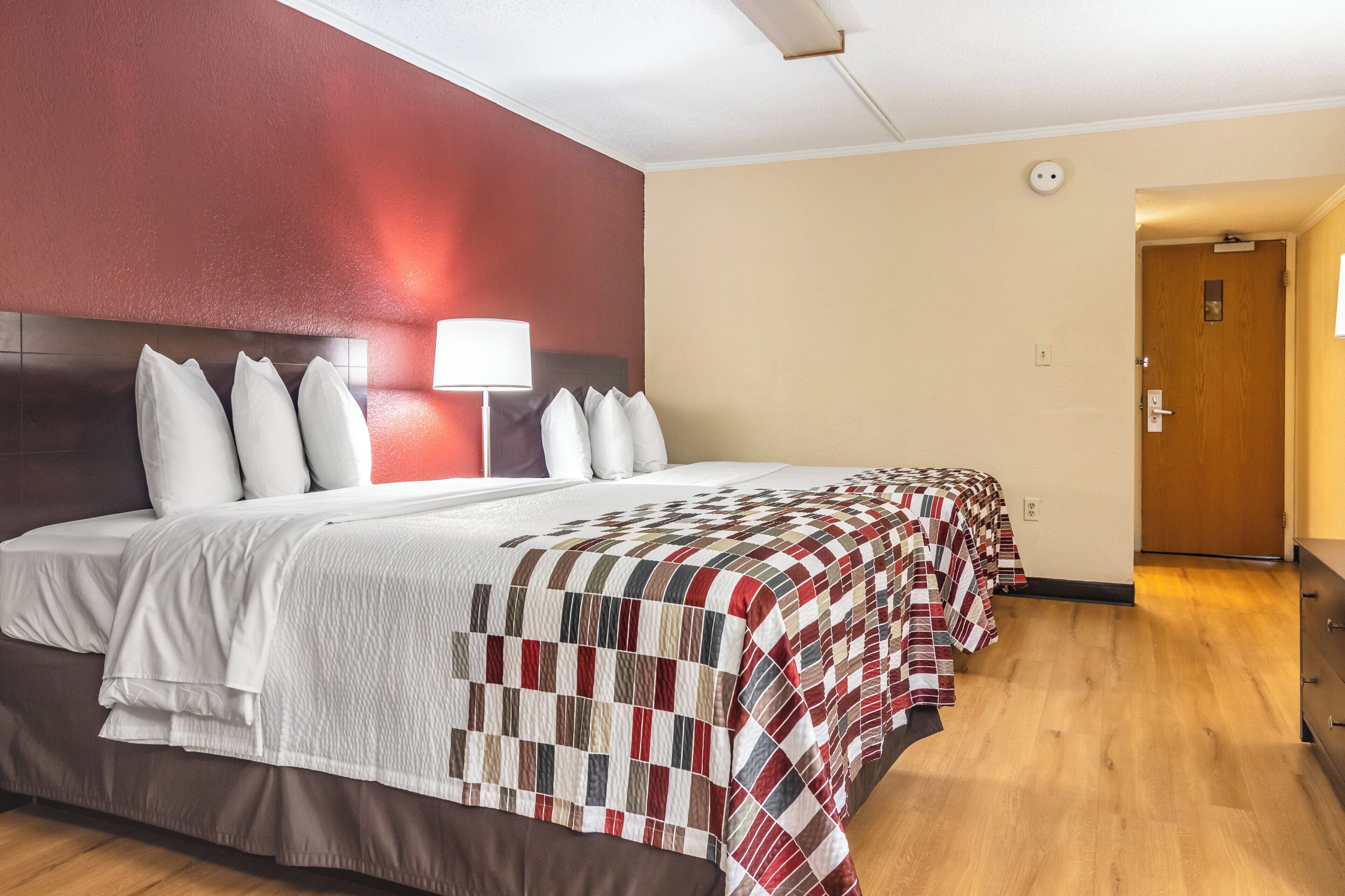 A hotel room with a large bed, a bedside lamp, a wooden floor, and a door to the right.